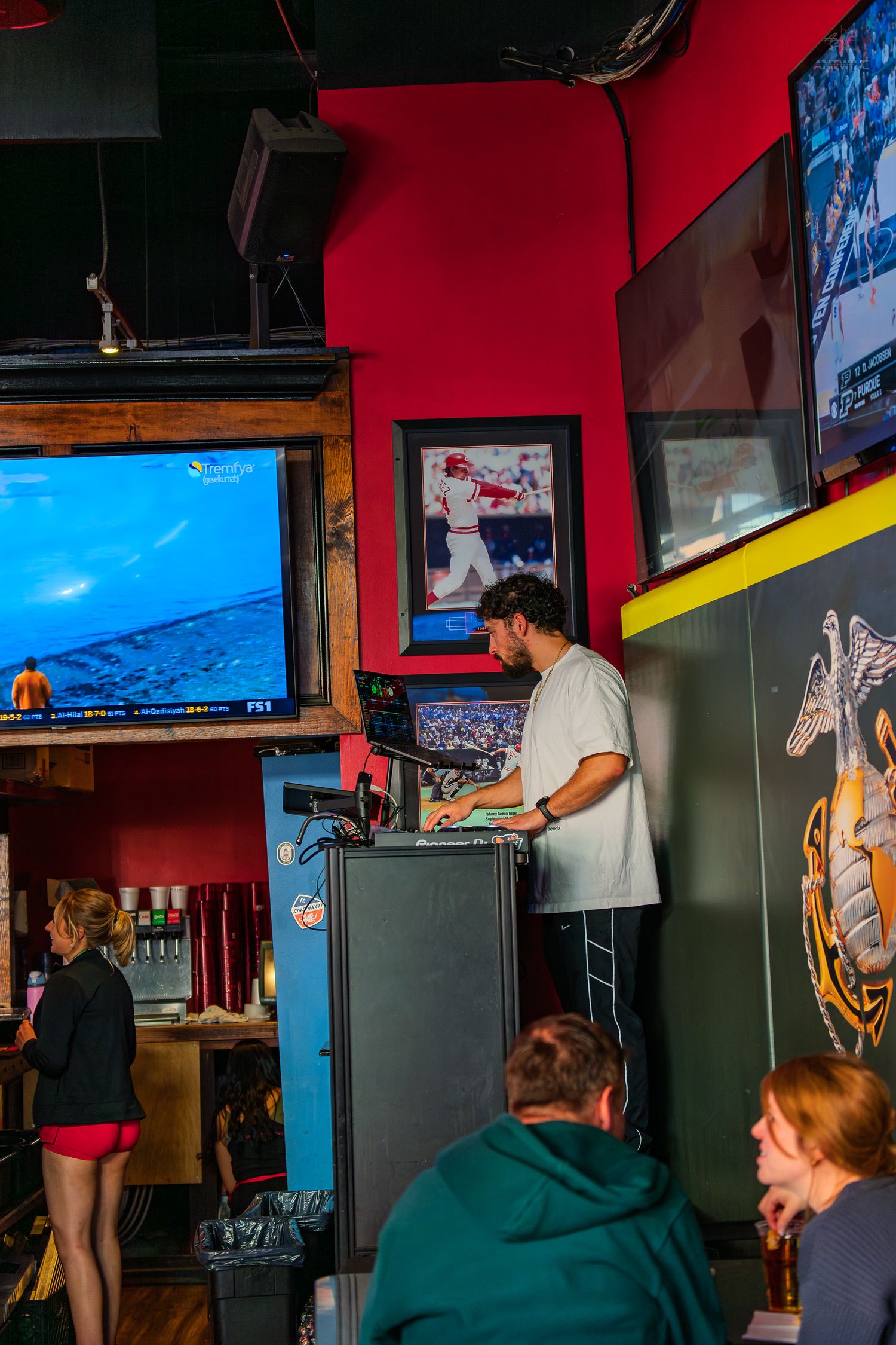 Man operates DJ booth in a sports bar with TVs showing baseball, while patrons socialize nearby.