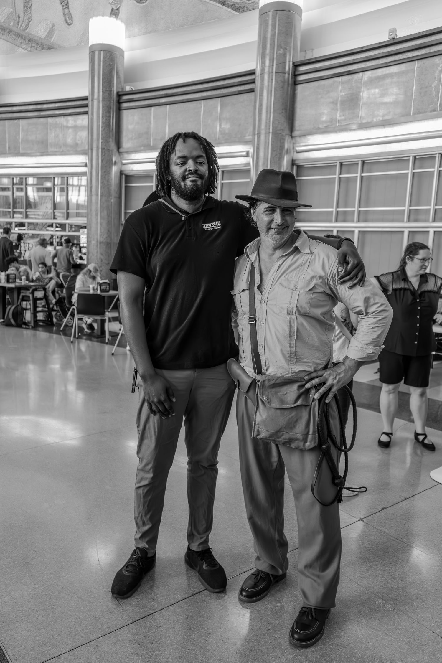Two men pose together in a convention hall, one in casual wear and one in an Indiana Jones costume.