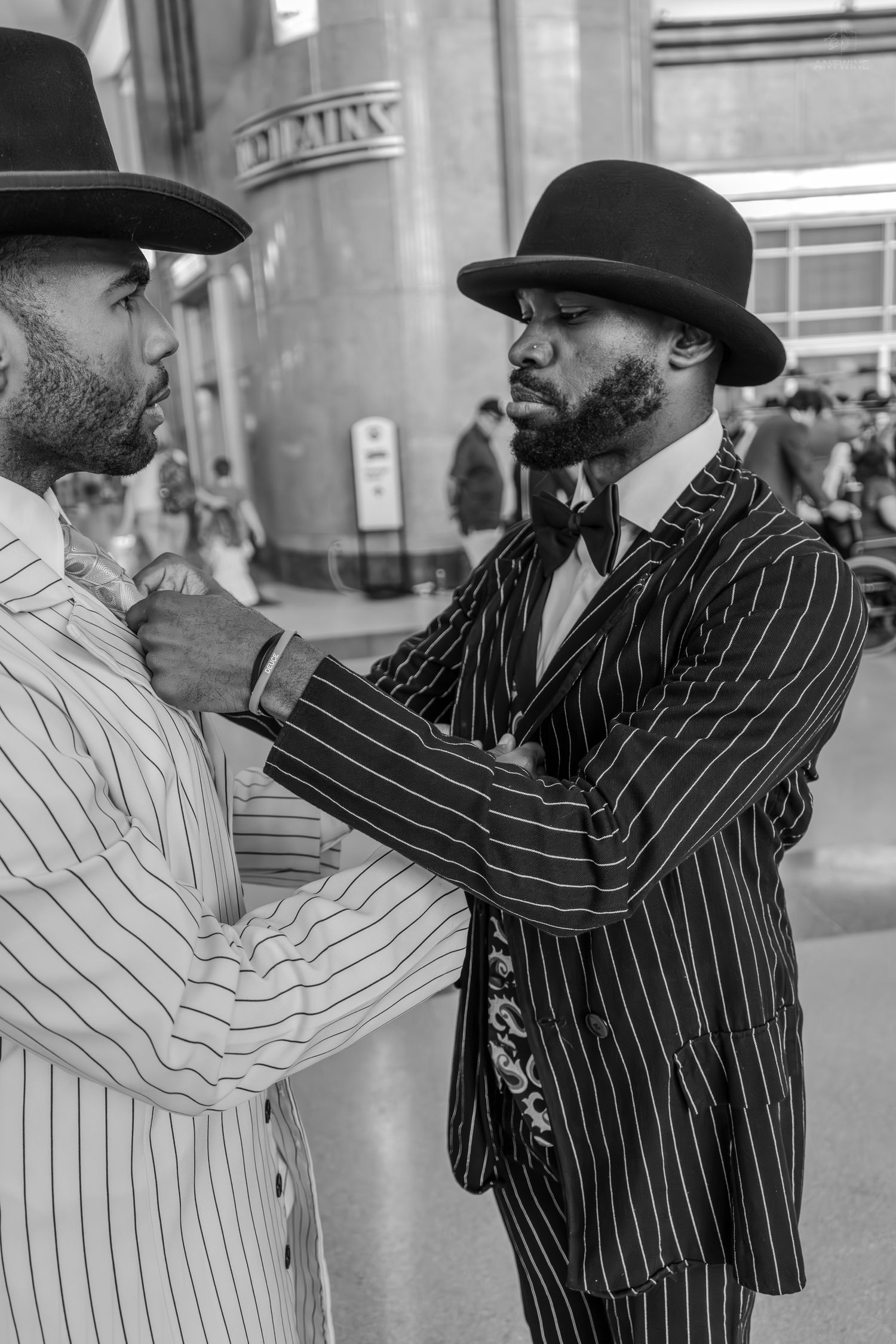 Two Black men in pinstripe suits and bowler hats interact on a city street in a stylish black and white photo.