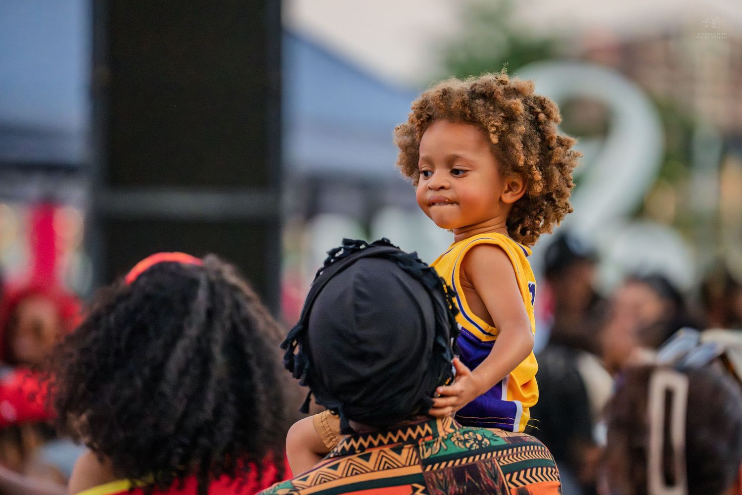 Adorable curly-haired toddler sitting on shoulders of adult at a vibrant Juneteenth block party.