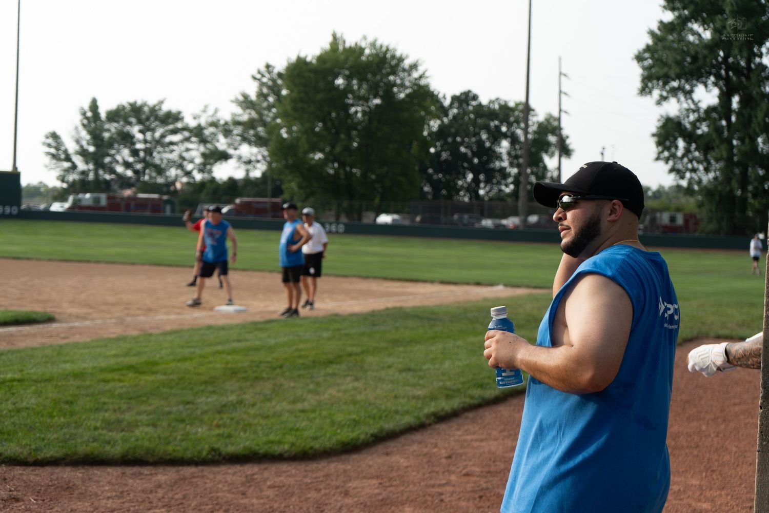 Man in blue sleeveless shirt and sunglasses holds drink at outdoor baseball field with players in background.