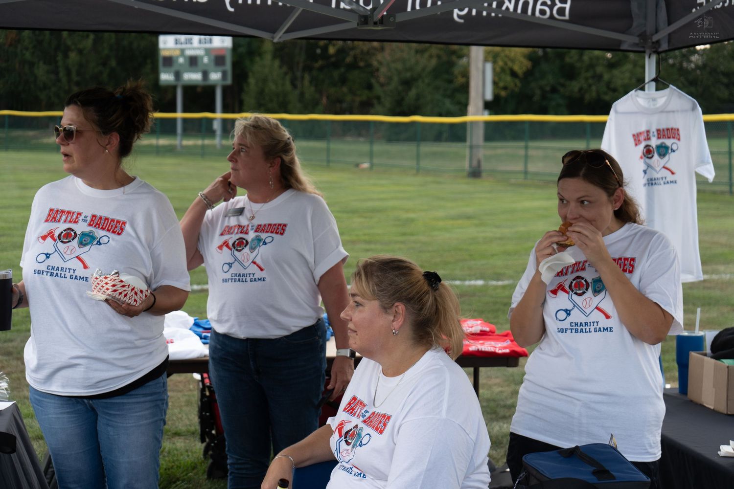 Four women in matching event t-shirts gather under a tent at an outdoor softball tournament.