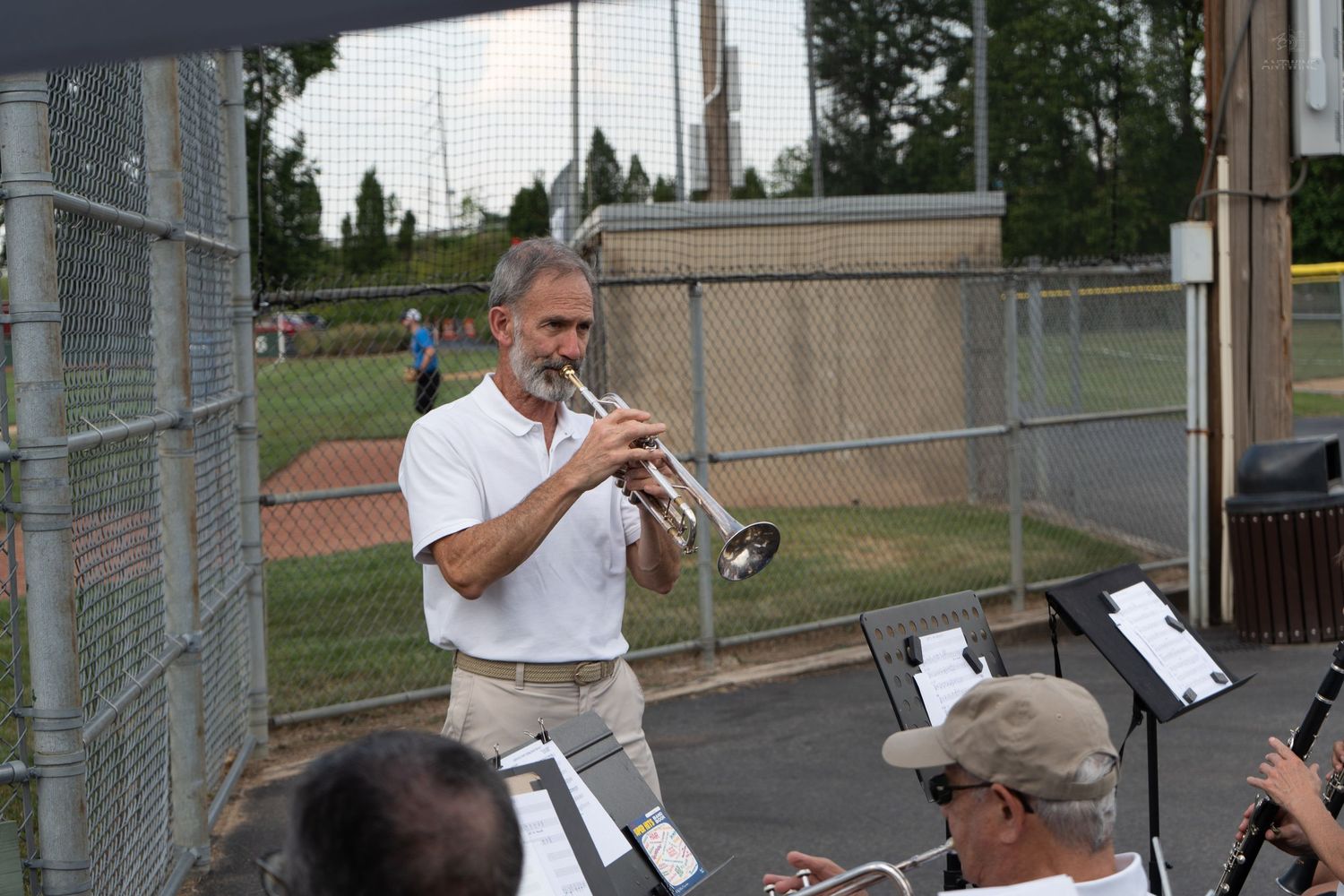 Man playing trumpet outdoors at a baseball field during an evening band performance with musicians in foreground.