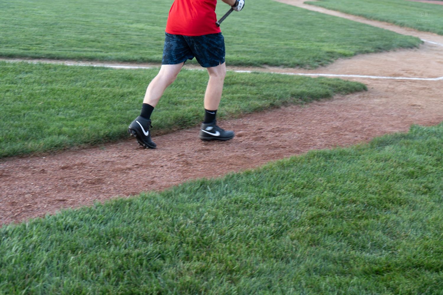 Close-up of baseball player in red shirt and black shorts running bases in Nike cleats on a grass baseball field.