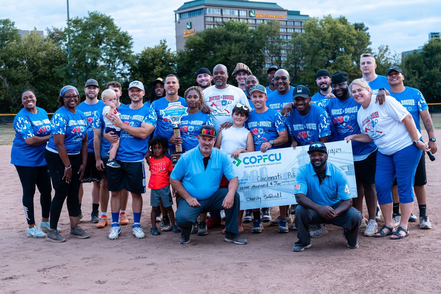 Large group of softball players and fans pose with a COPFCU ceremonial check on a baseball field after a charity game.
