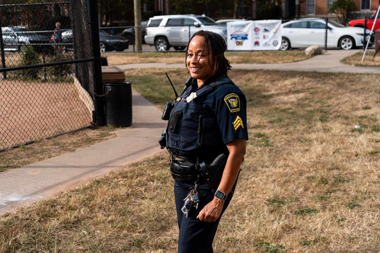 Female police sergeant in uniform standing outdoors on a sunny day near a chain-link fence.