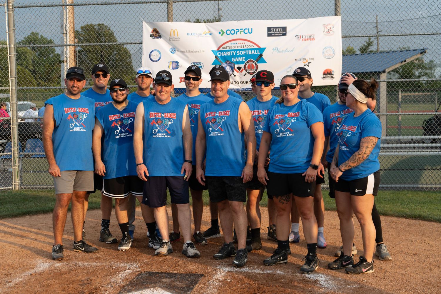 Softball team posing together in matching blue shirts on a baseball diamond at an outdoor tournament.