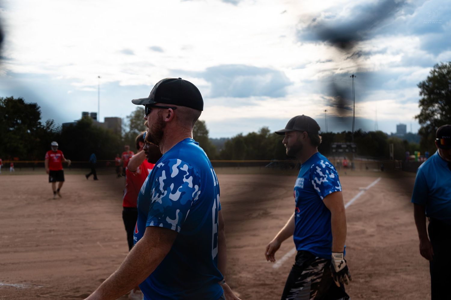 Men in blue team jerseys walk on a softball field under cloudy skies during an outdoor recreational game.