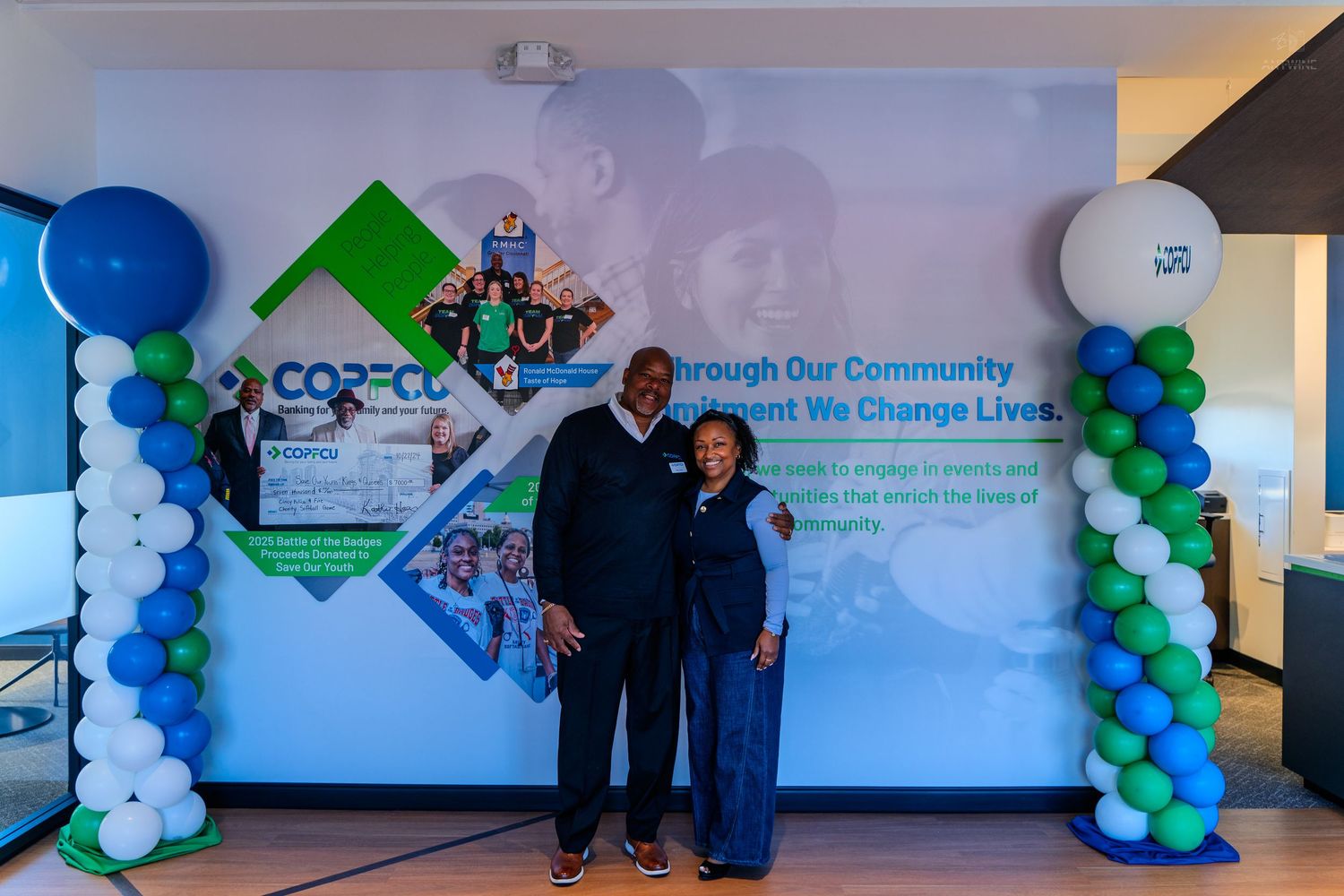 Two people pose together at a COFCU community event, surrounded by blue and green balloon columns and a branded backdrop.