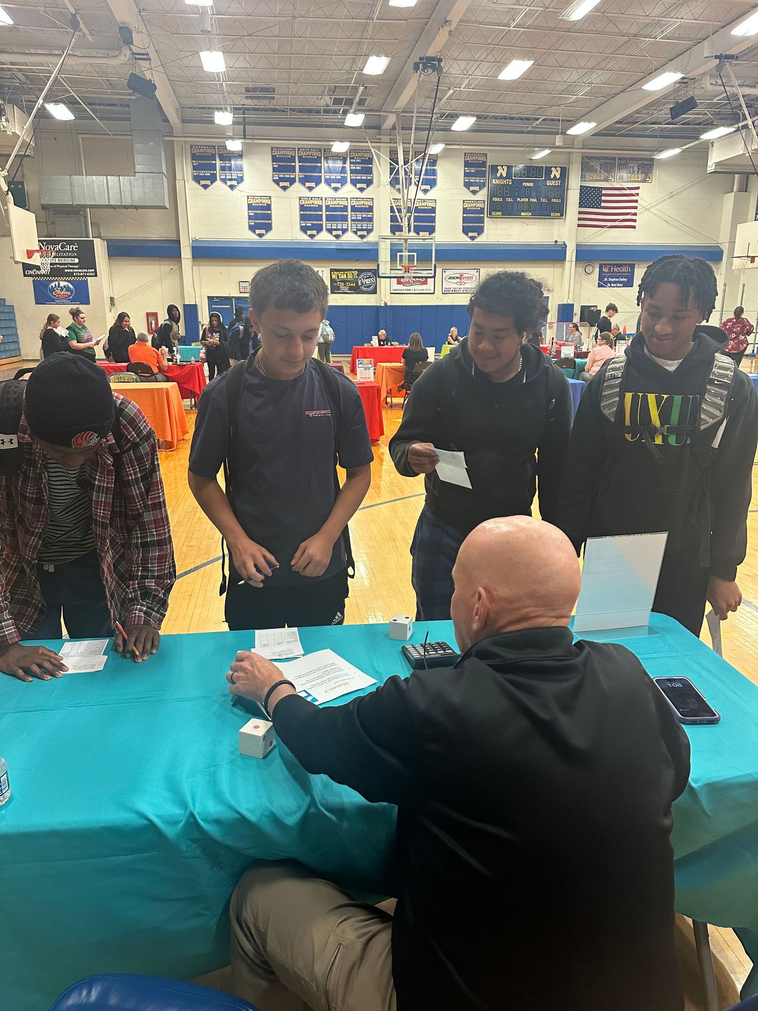 Students line up at a teal table in a school gymnasium for a college fair or registration event.