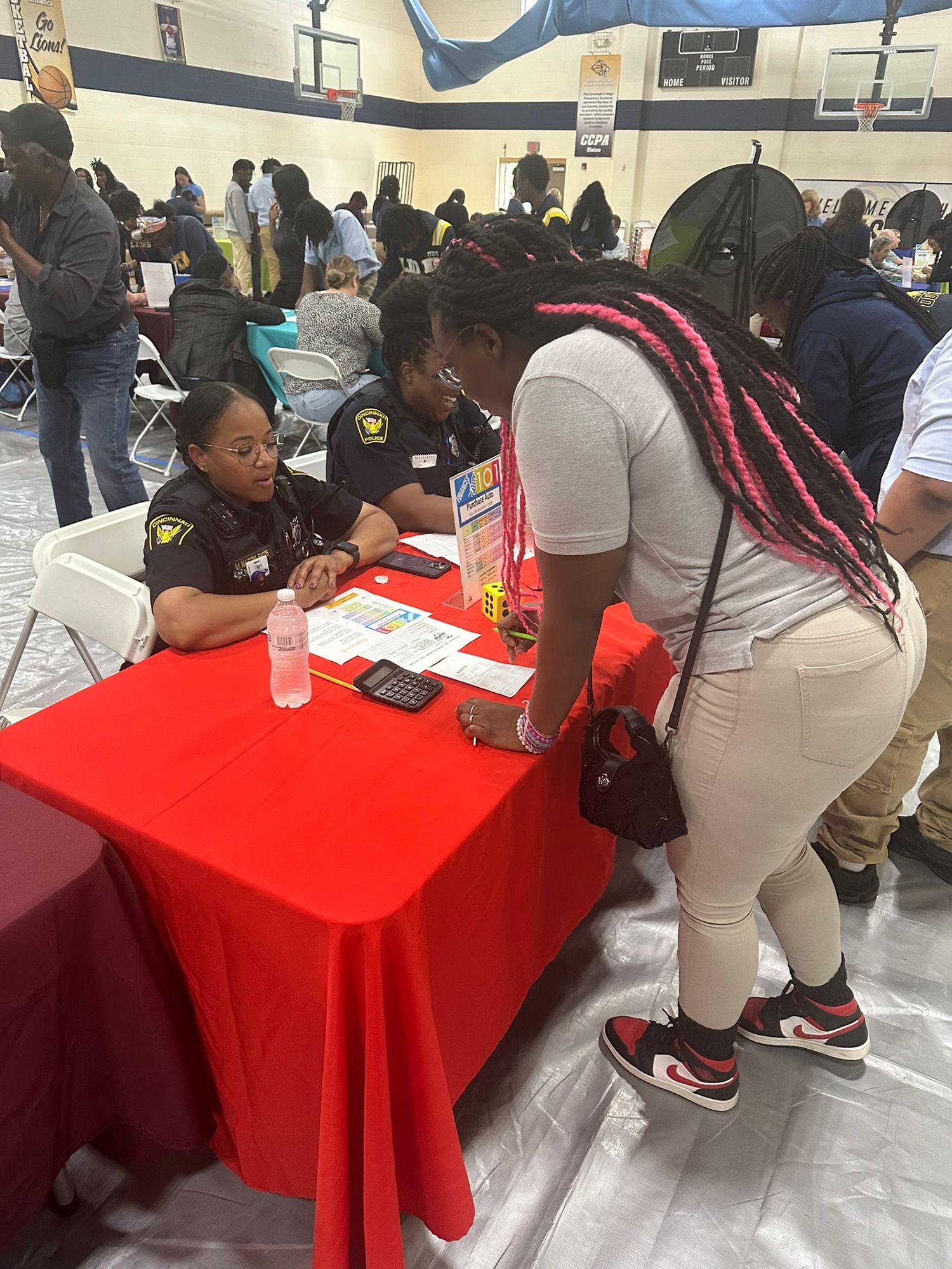 Two officers in uniform speak with a young woman at a red table during an indoor career fair event.