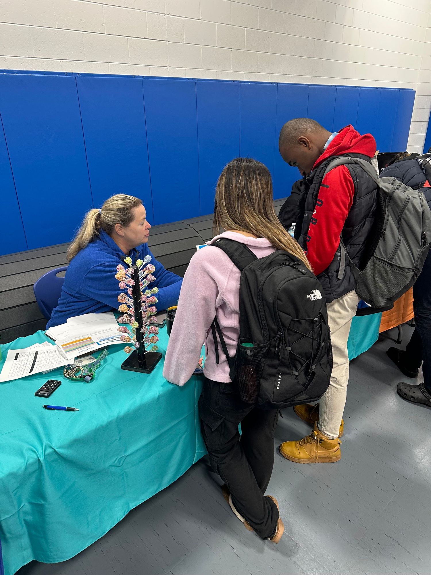 Students with backpacks speak with a female recruiter at a teal table during a school career fair.