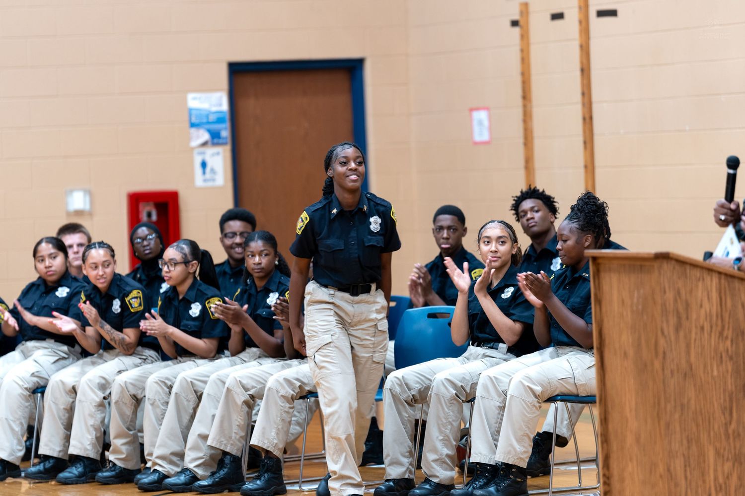 Young students in matching police cadet uniforms clap and cheer during an indoor assembly ceremony.