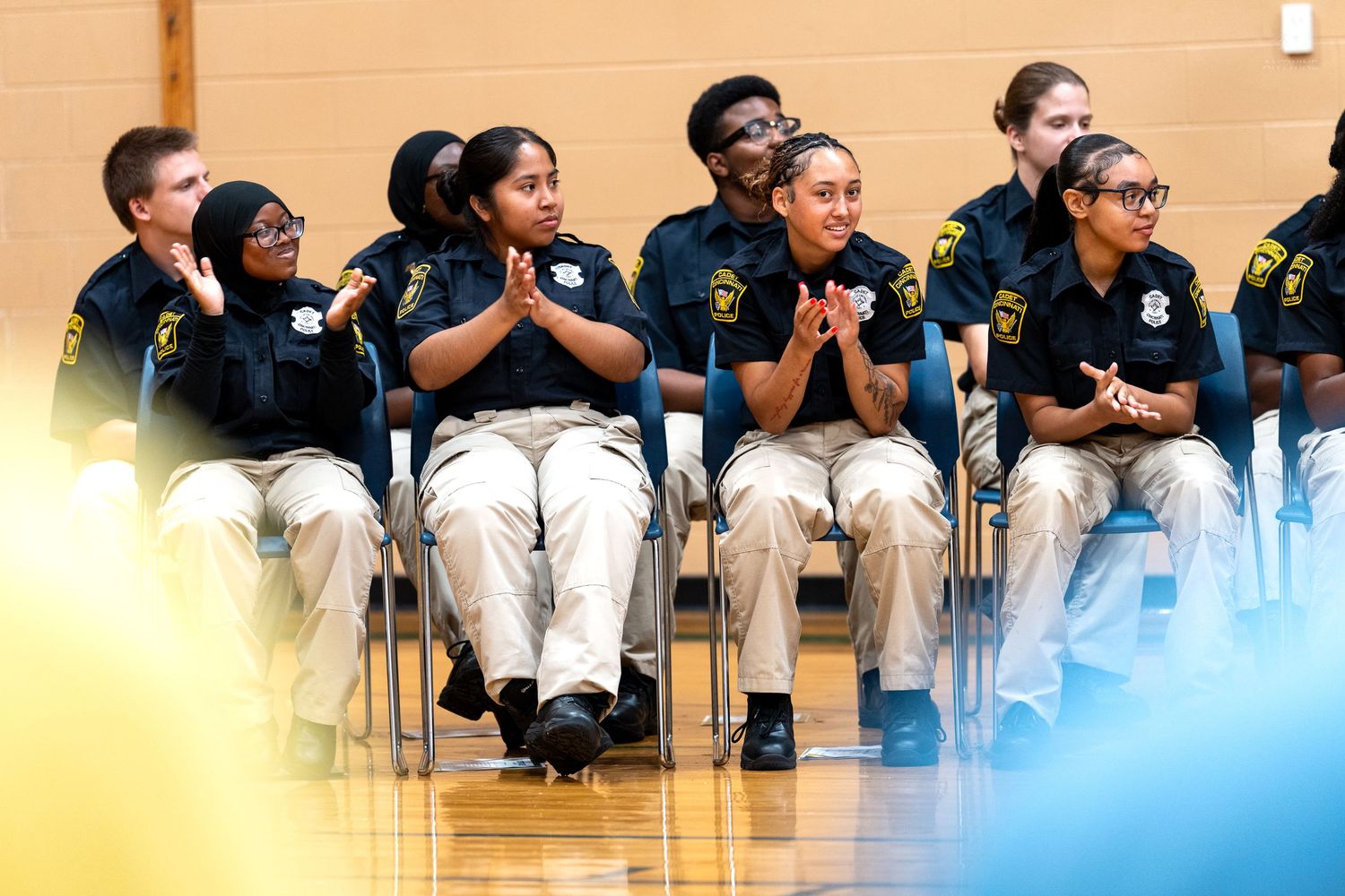 Young cadets in uniform shirts and khaki pants sit clapping at an indoor ceremony or graduation event.