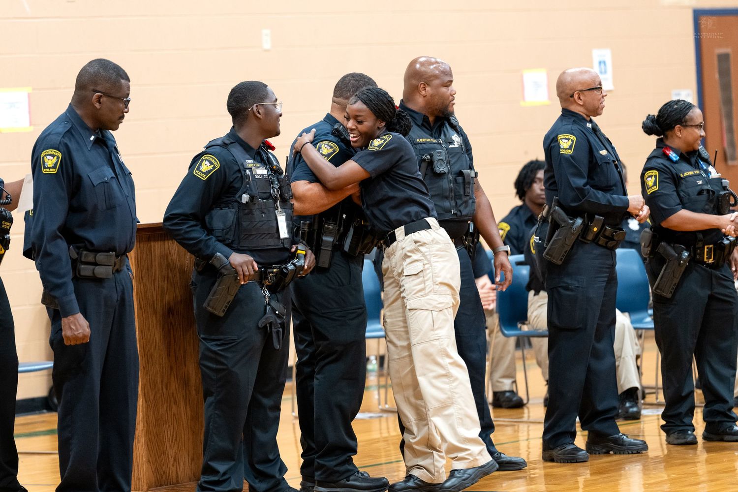 Law enforcement officers in uniform share an emotional embrace during a formal ceremony in a gymnasium.