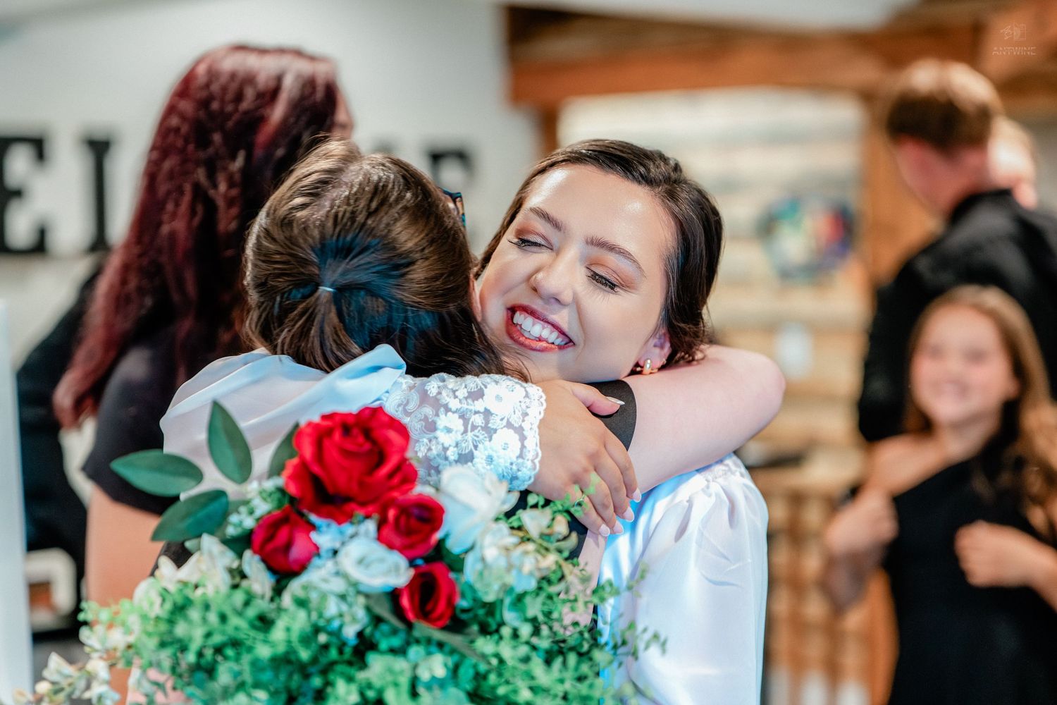 Bride smiles radiantly while embracing a guest holding red roses at wedding reception.