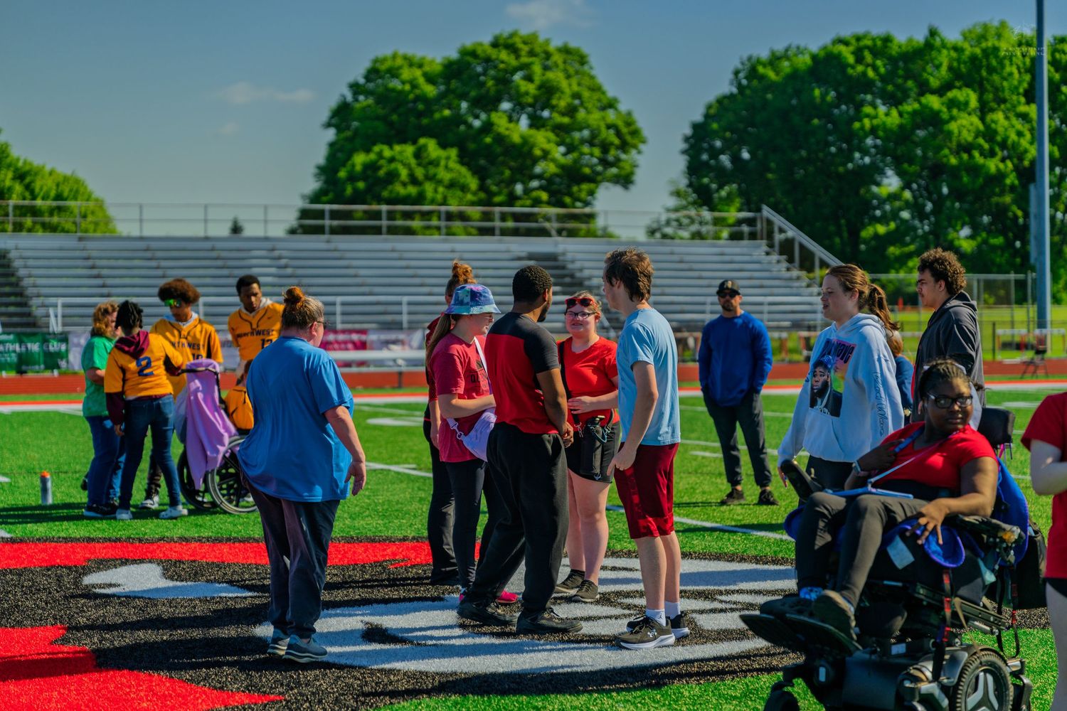 Students gather on a red track at an outdoor stadium during a sunny school sports event surrounded by green trees.