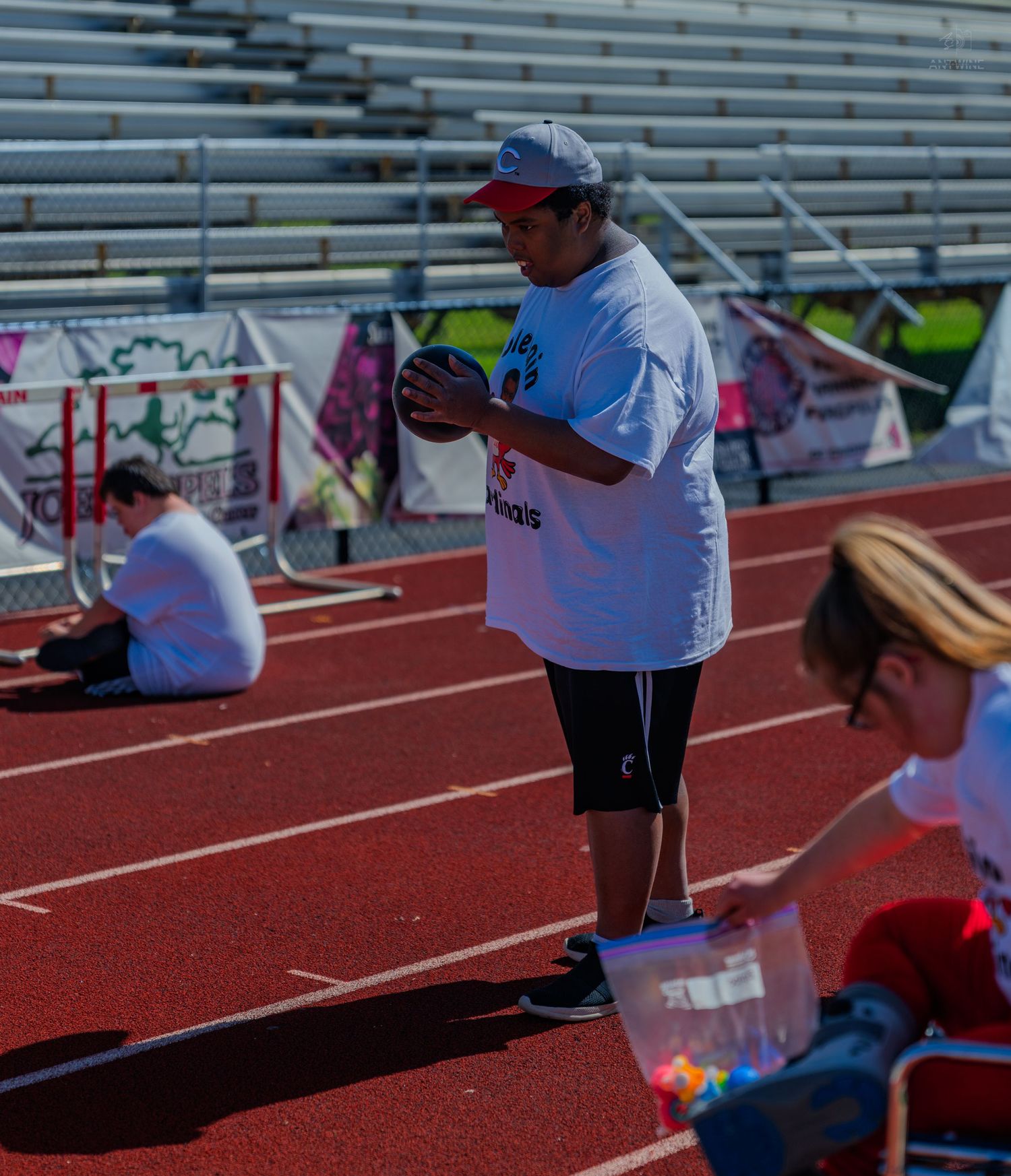 Man in white t-shirt and cap holds a ball on a red running track near stadium bleachers during an outdoor event.