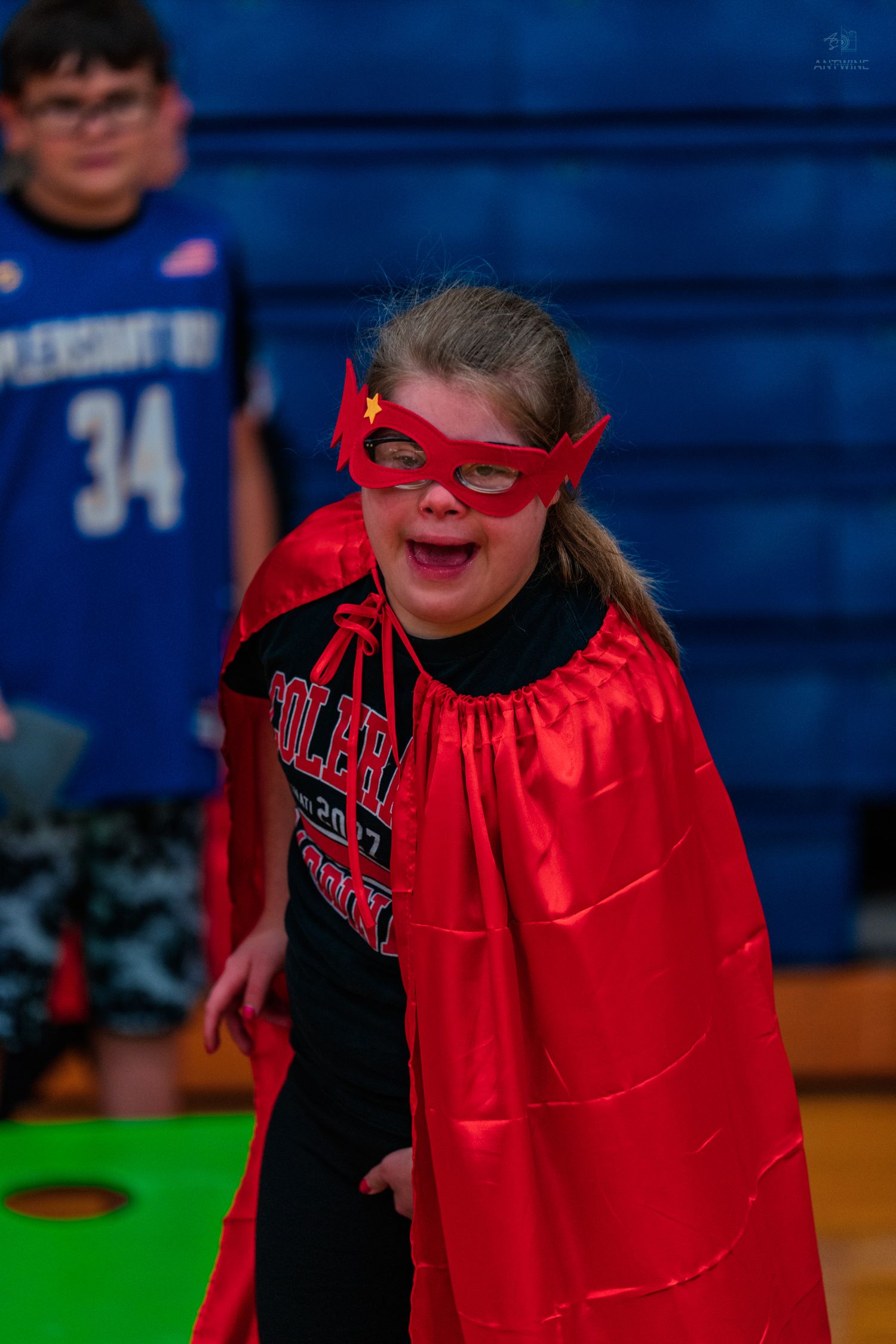 Young girl wearing red superhero cape and mask smiling joyfully in a school gymnasium.