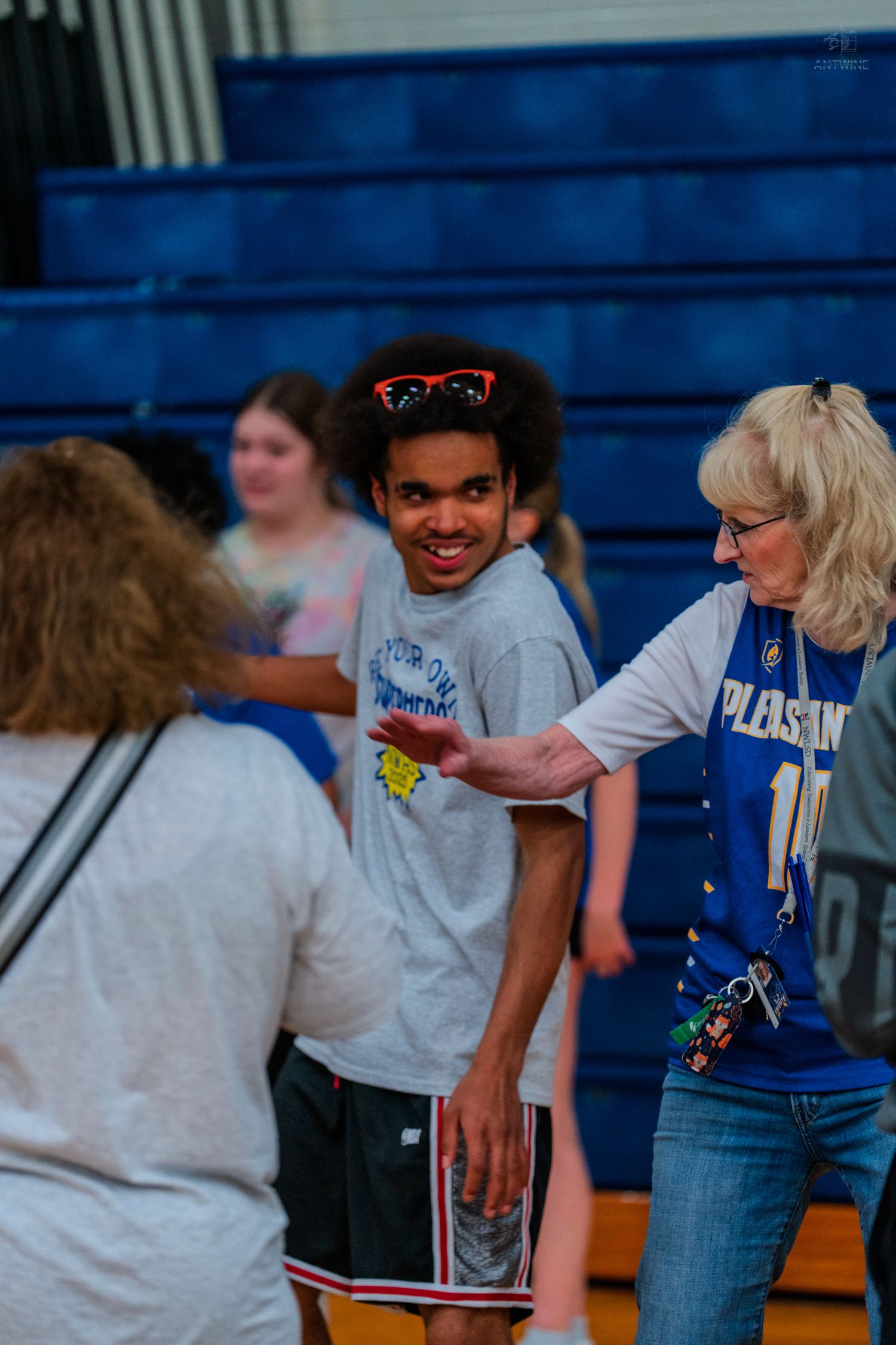 Students interact with a teacher in a school gymnasium, with blue bleachers visible in the background.