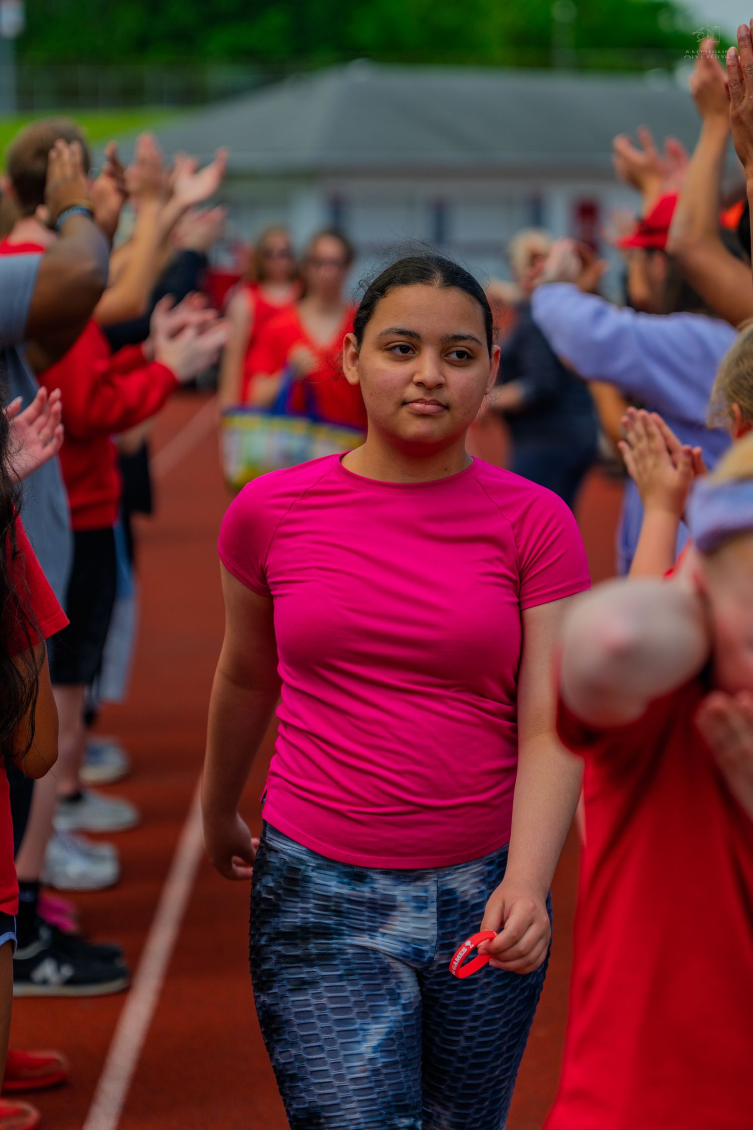 A girl in a pink shirt walks through a cheering crowd on a red track during a school sports event.