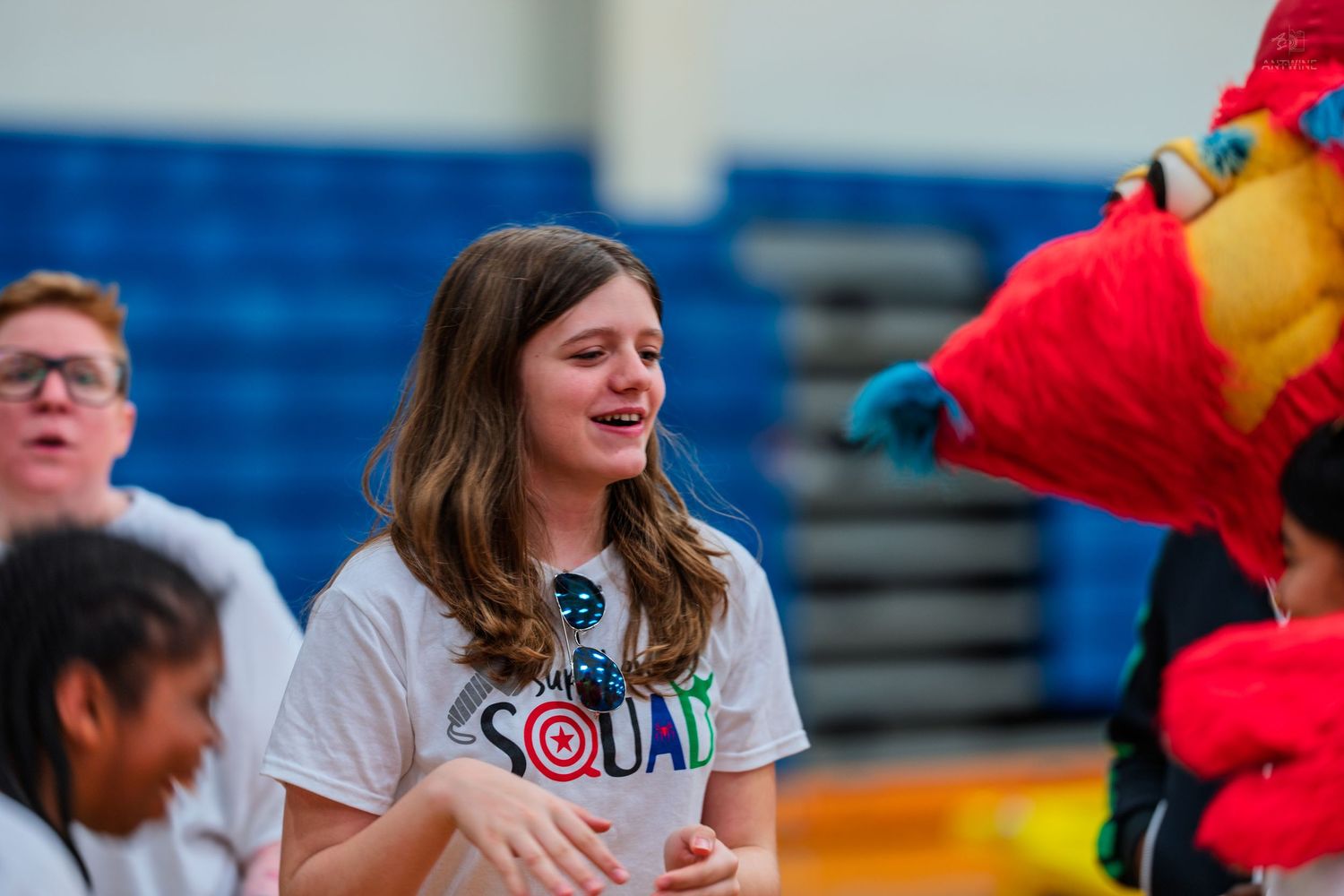 A smiling girl in a Squad t-shirt interacts with a colorful mascot at a school gymnasium event.
