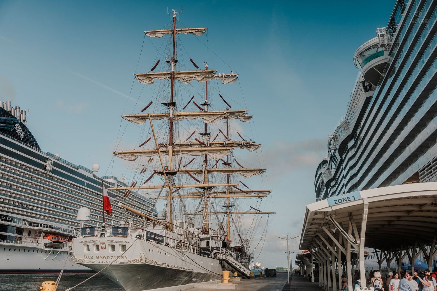 A classic tall ship with full masts docked beside a modern cruise ship at a sunny Caribbean port terminal.