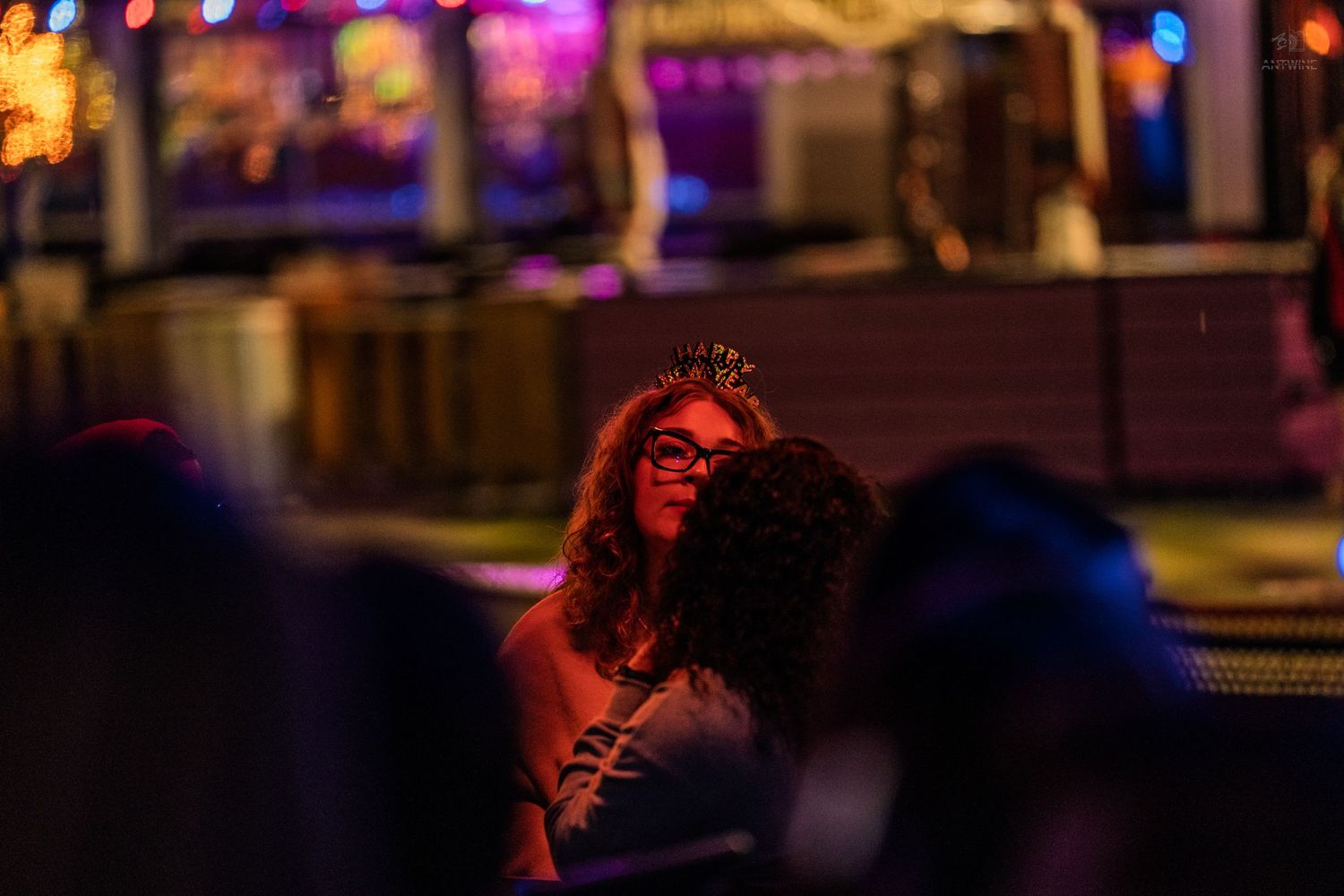 Woman wearing a tiara and glasses relaxing in a dimly lit bar with colorful neon lights.
