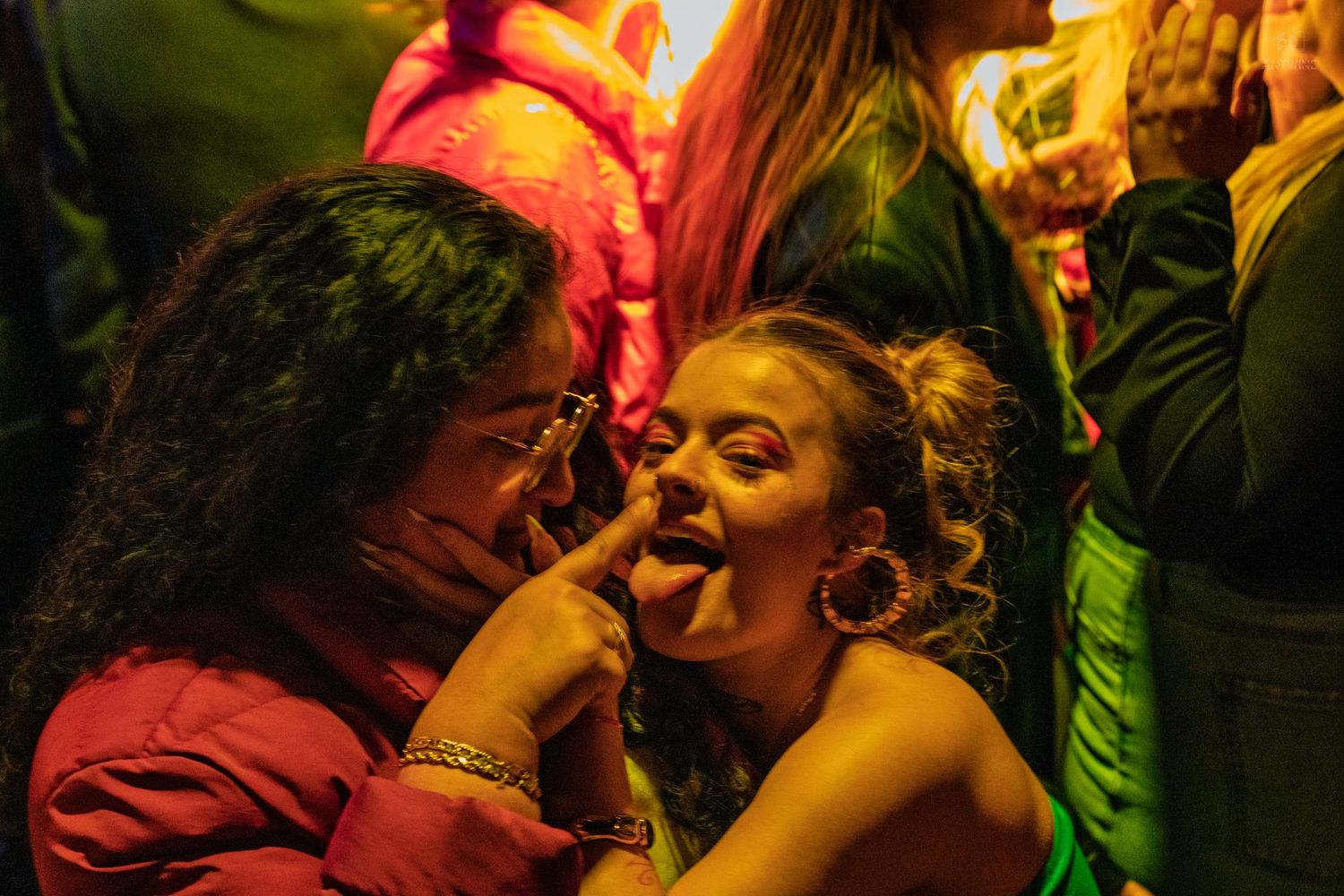 Two women laugh together at a vibrant nightclub, one playfully sticking out her tongue.