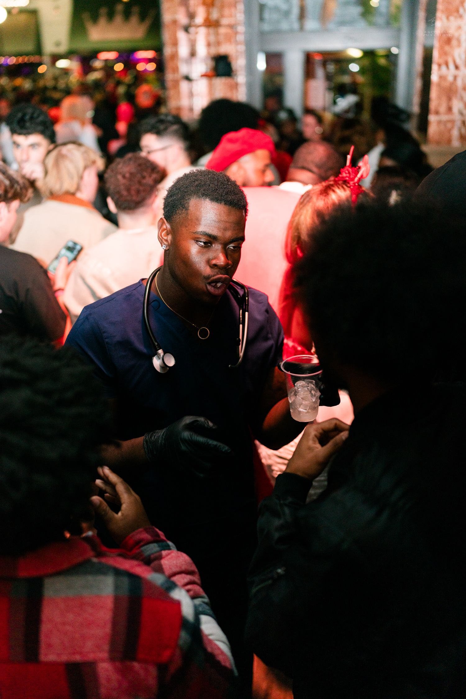 Young man in scrubs costume holding a drink and conversing at a packed Halloween party event.
