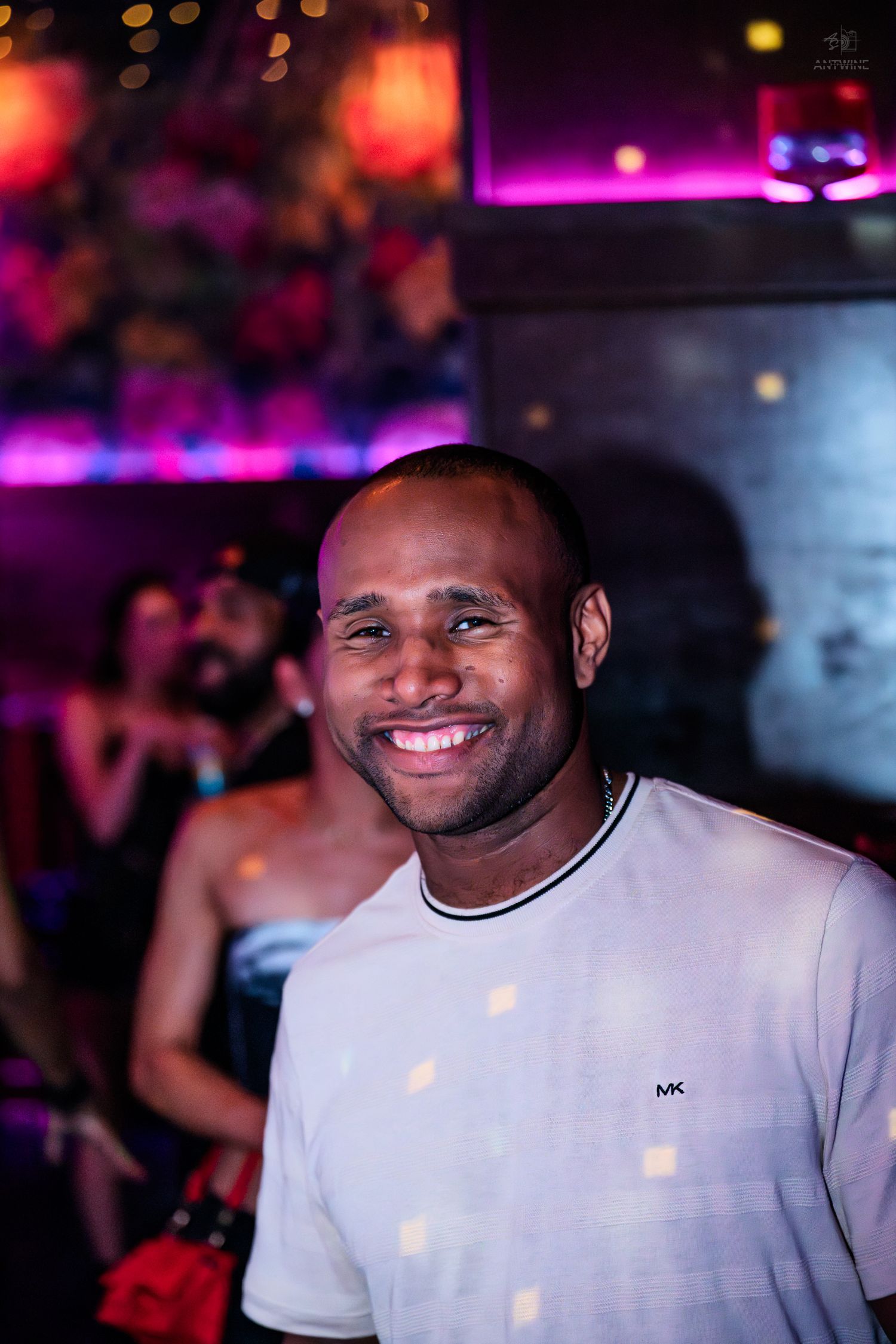 A smiling bald Black man in a Michael Kors cream t-shirt poses in a neon-lit nightclub.