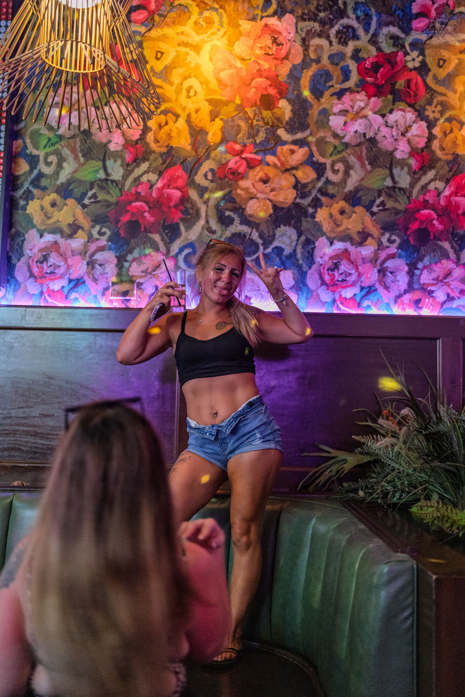 Confident woman in crop top holding drink poses before colorful floral mural in nightclub.