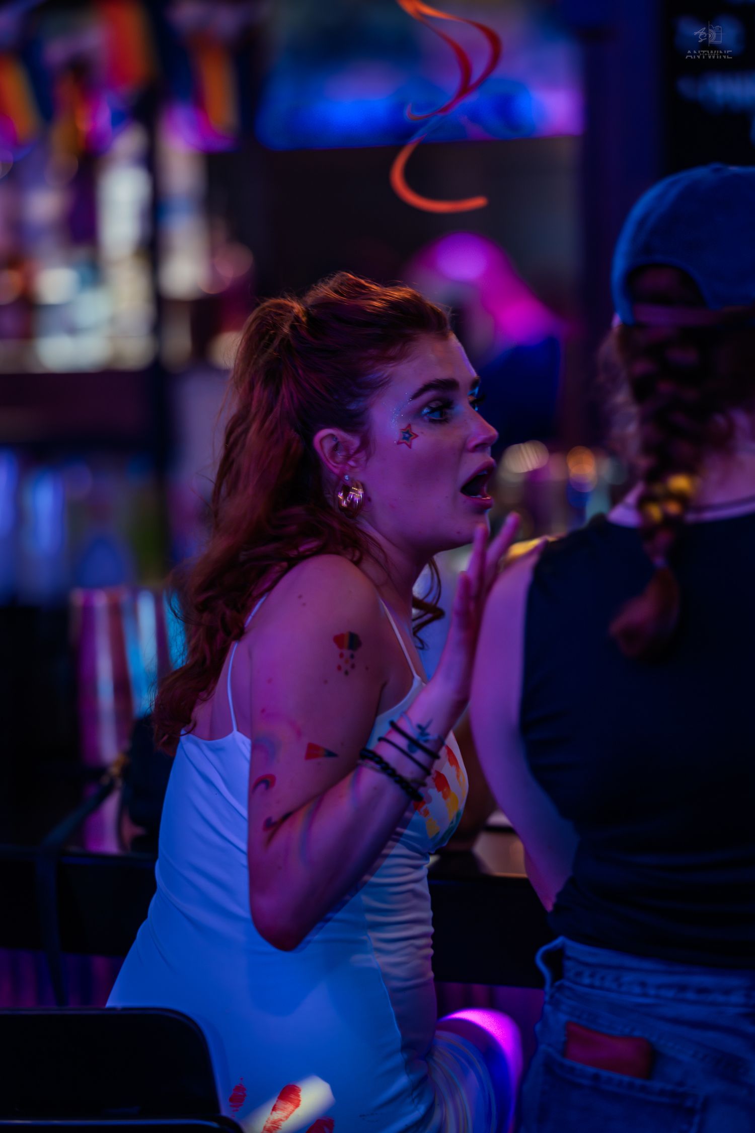 A redhead woman with face stickers reacts expressively at a colorful neon-lit nightclub.