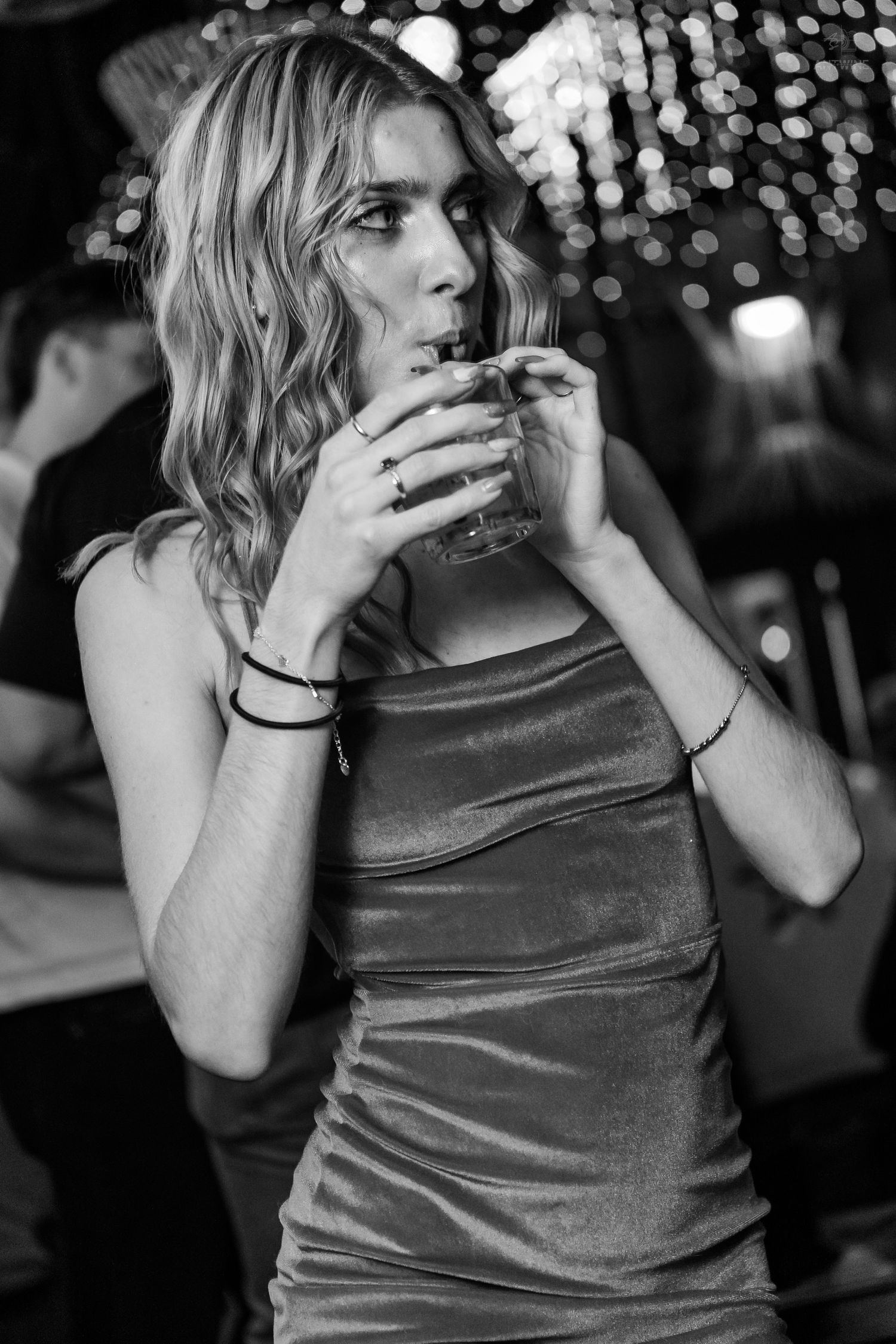 Blonde woman in silver velvet dress drinking through a straw at a nightclub.