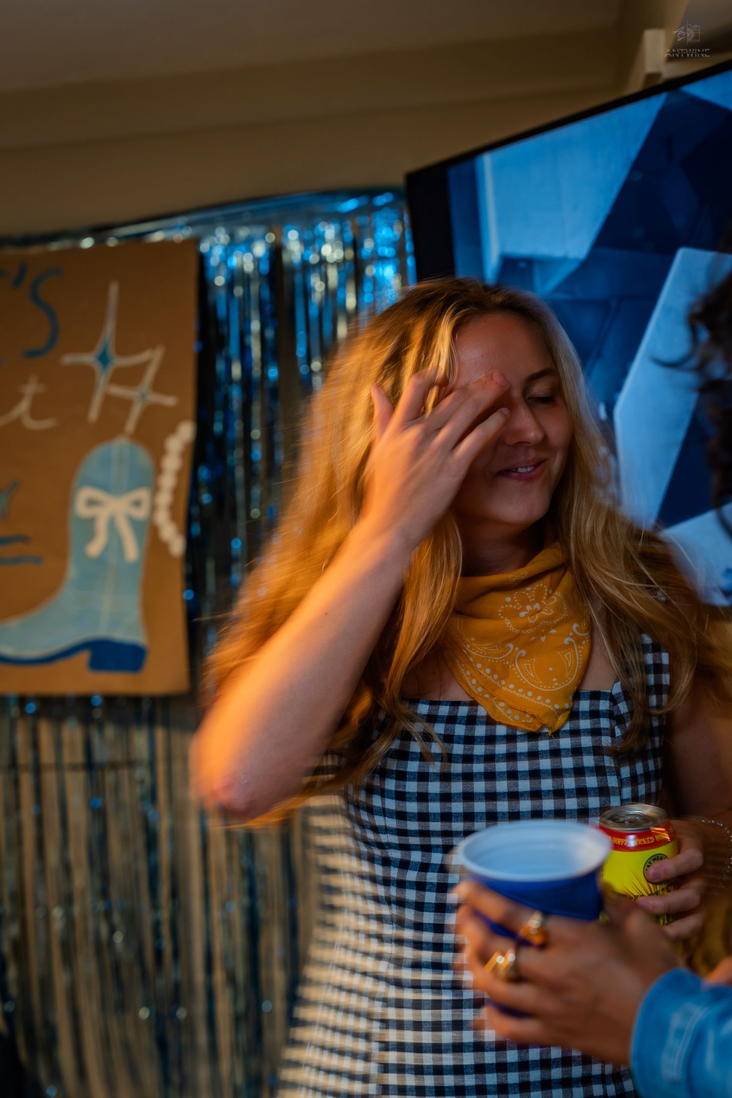 Laughing woman in gingham dress with yellow bandana holding cup at a birthday party celebration.