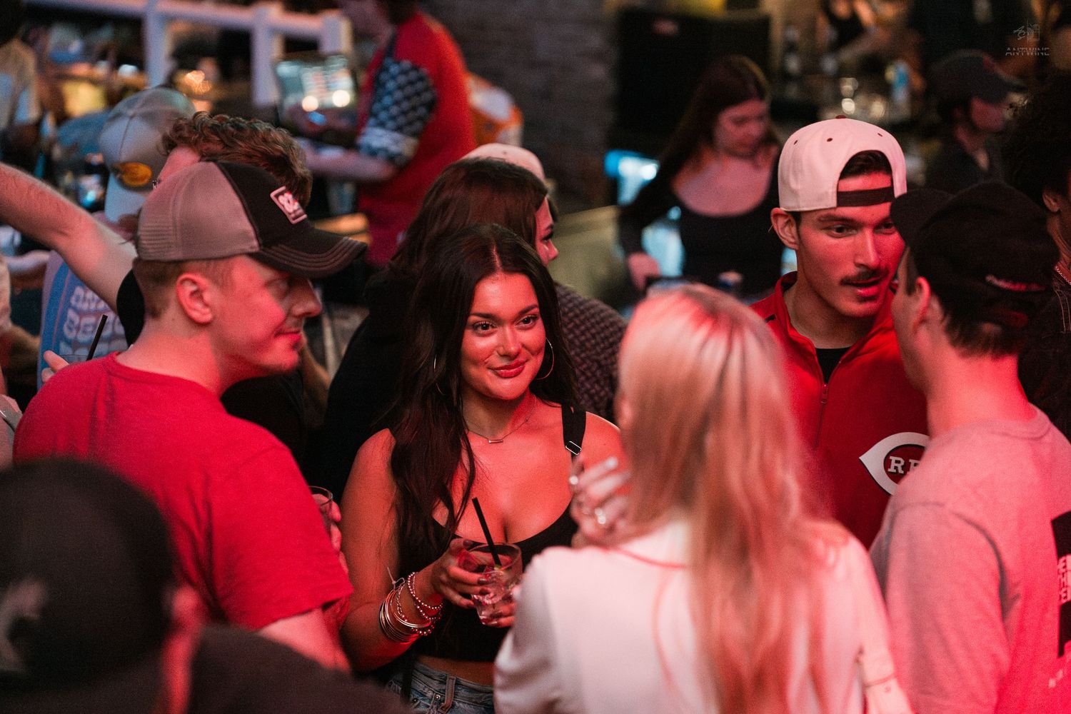 A group of smiling friends socializing under red lights at a crowded Cincinnati bar night.