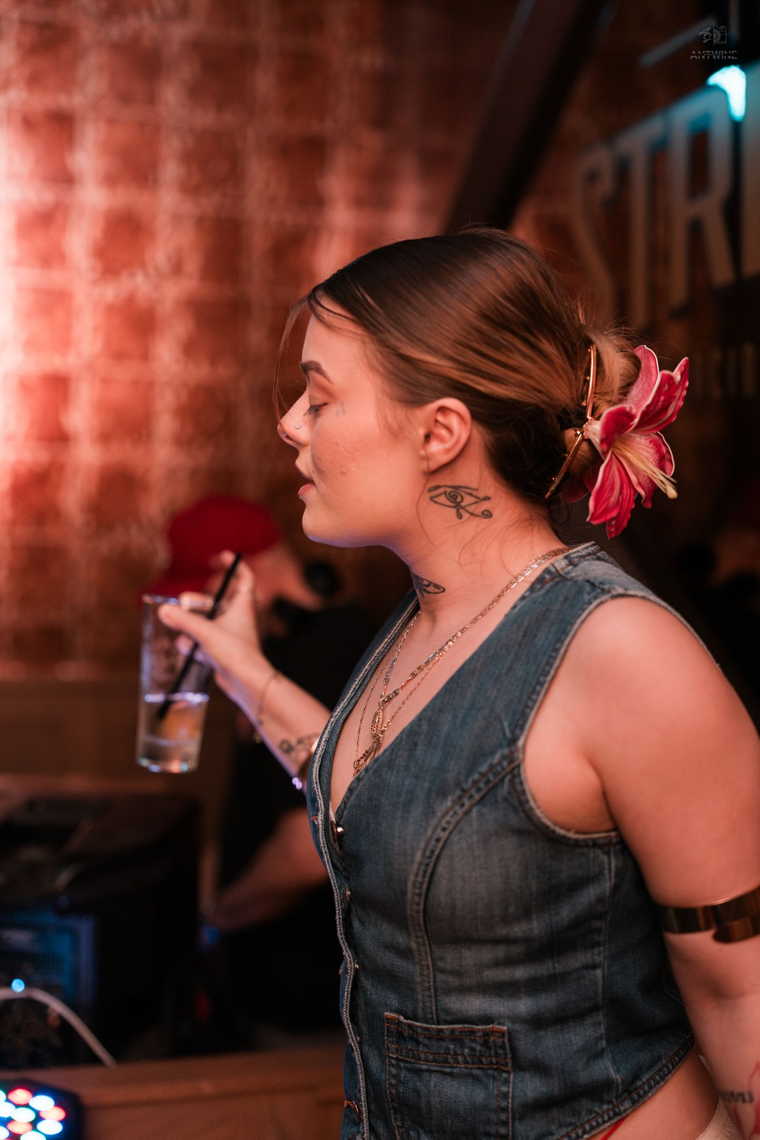 Young woman with flower hair accessory and neck tattoos sipping a drink at a dimly lit bar.