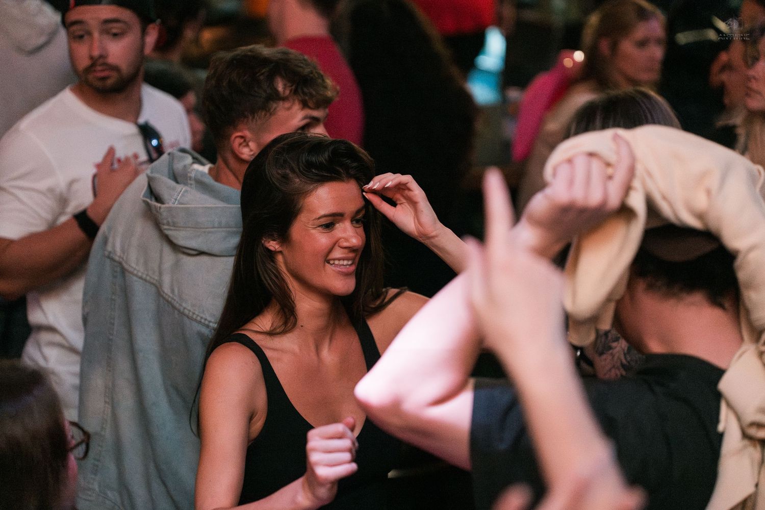 Young woman in black top dancing and smiling at a crowded nightclub party event.