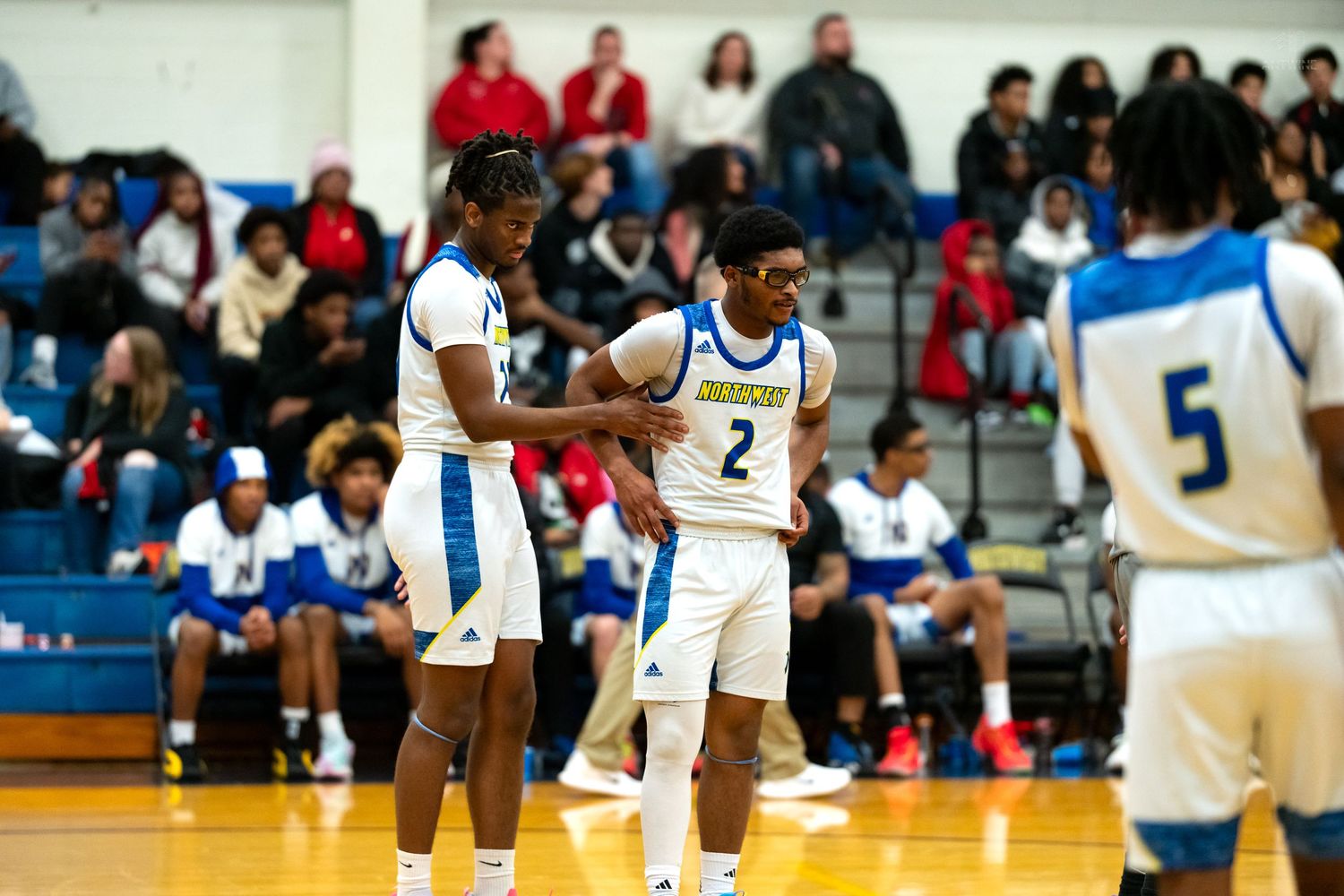 Two basketball players in white and blue uniforms stand together on an indoor court during a high school game.