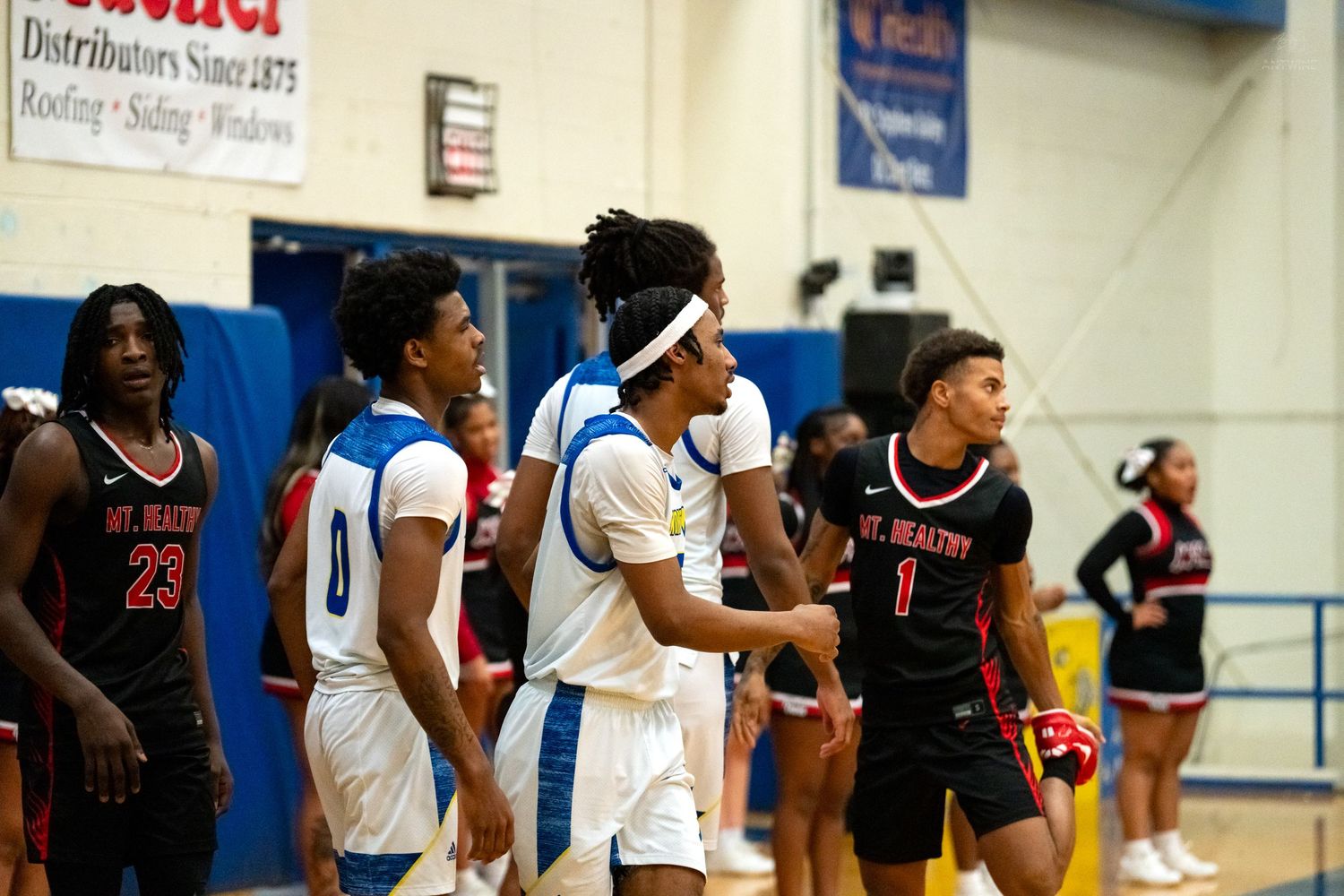 High school basketball players from two teams stand near mid-court during a pause in a gymnasium game.