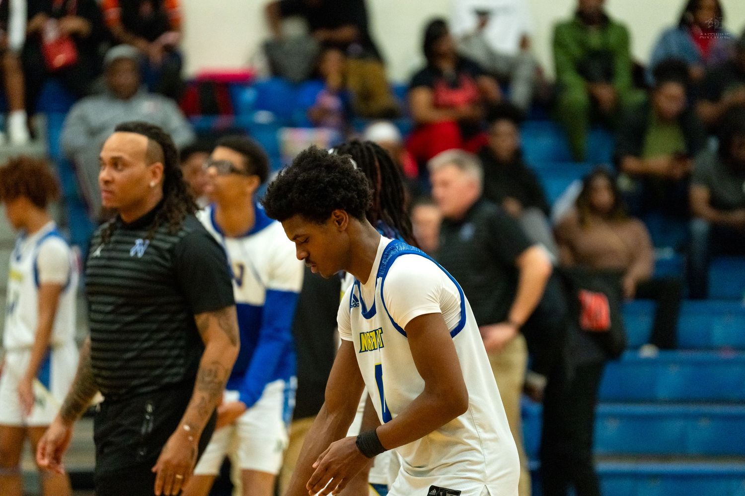 A high school basketball player in blue and white uniform walks on court during a game with coaches and crowd in background.