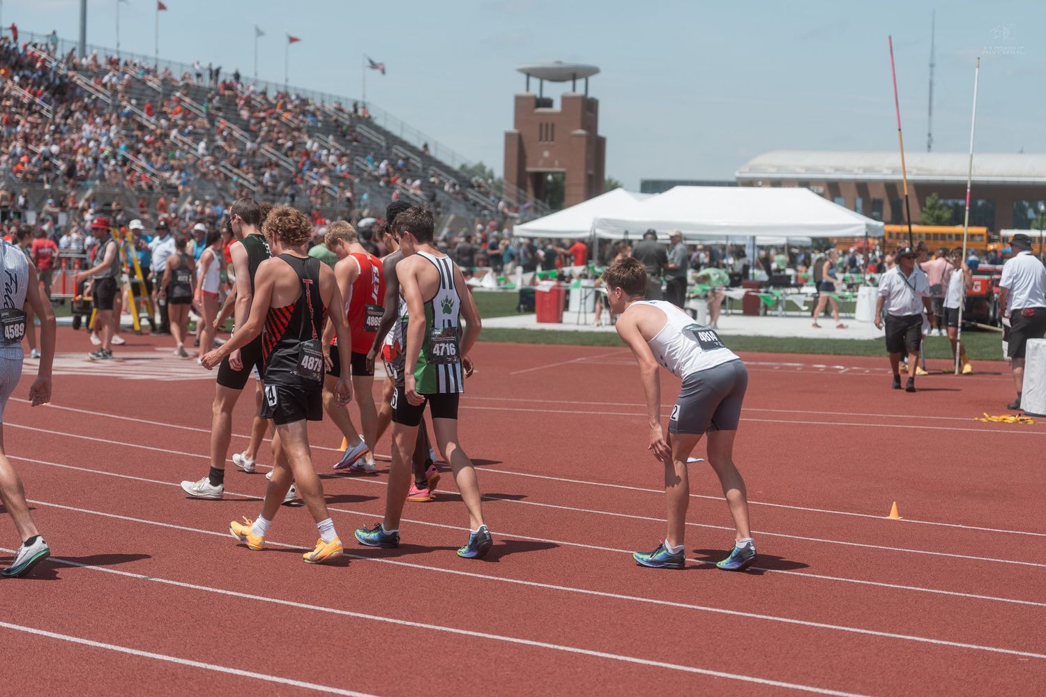Track and field athletes gather near the starting area on a red track at a crowded outdoor stadium.