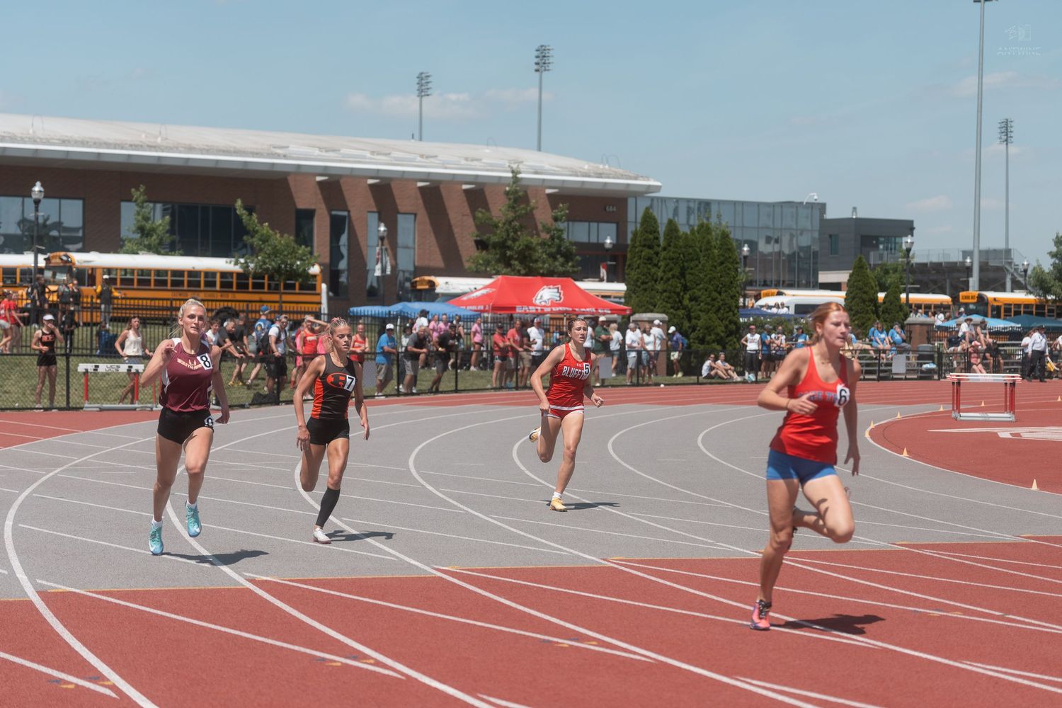 Female athletes racing on a red outdoor track during a sunny high school track and field competition.