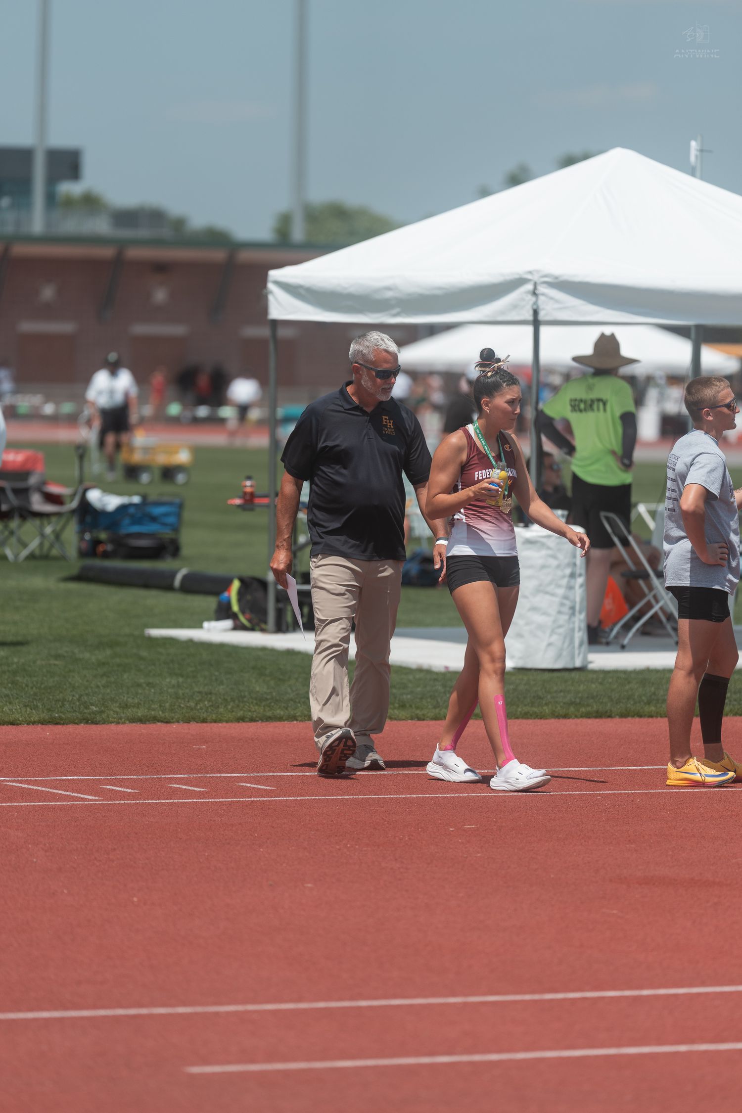 A coach walks alongside a female track athlete wearing a gold uniform at an outdoor sports event.