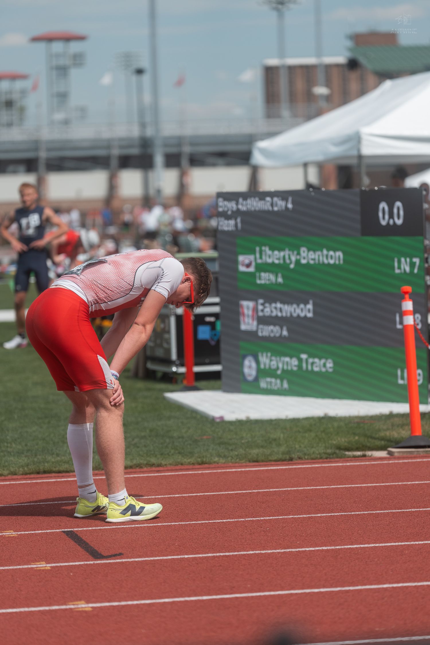 Exhausted male track athlete bends over catching breath near scoreboard showing Liberty-Benton results.