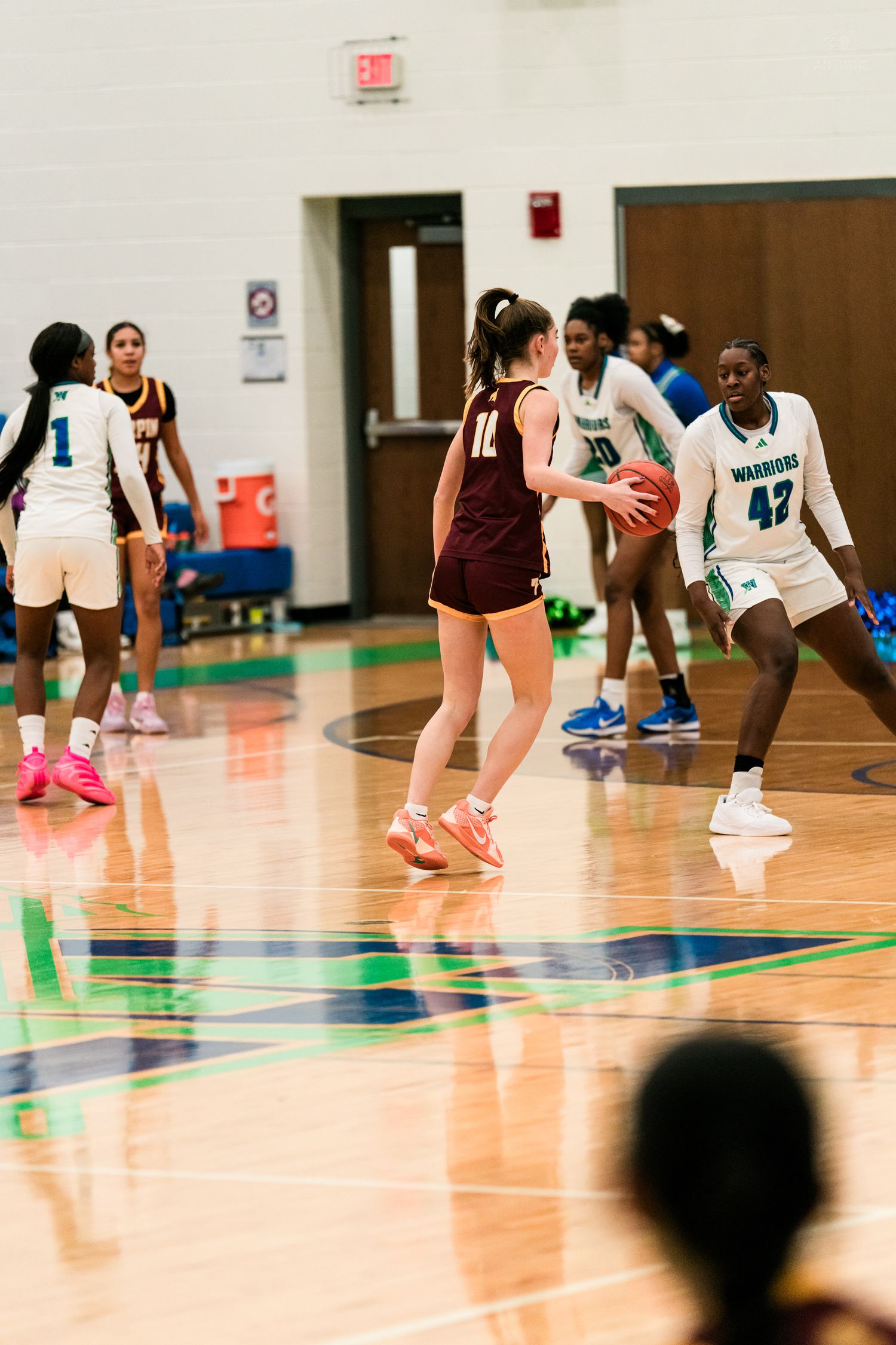 Female basketball players compete during a game, with a Turpin player in maroon holding the ball against Warriors defenders.