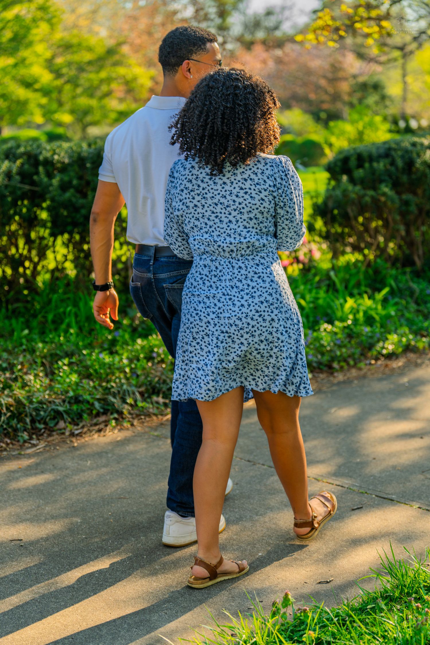 A couple walking hand-in-hand down a sunlit park path, surrounded by vibrant green hedges and trees.