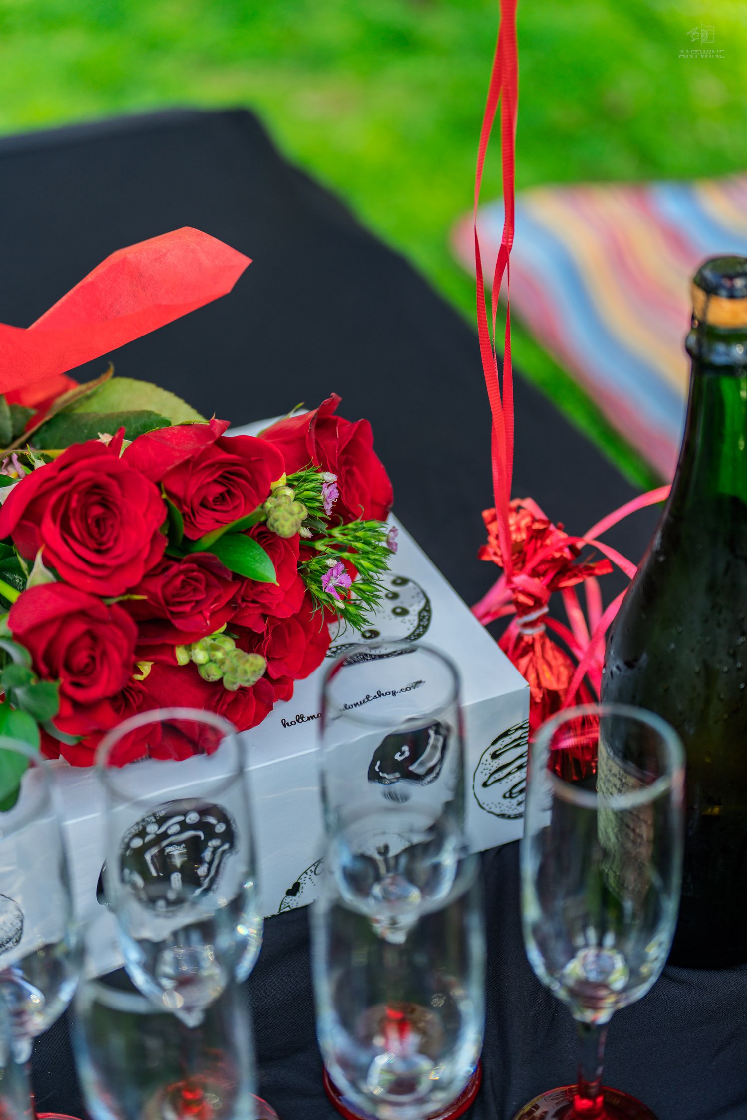 Red rose bouquet beside champagne flutes and a green bottle on a black table outdoors.