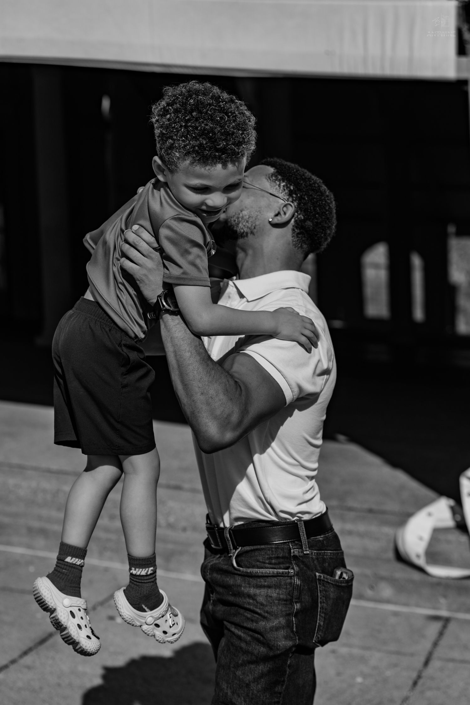 Black and white photo of a man lifting and kissing a young boy in the air outdoors.