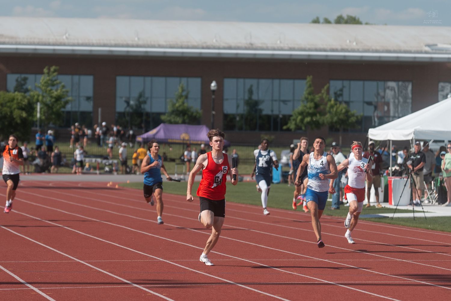 Male runner in red uniform leads competitors in a sprint race at an outdoor track and field meet.